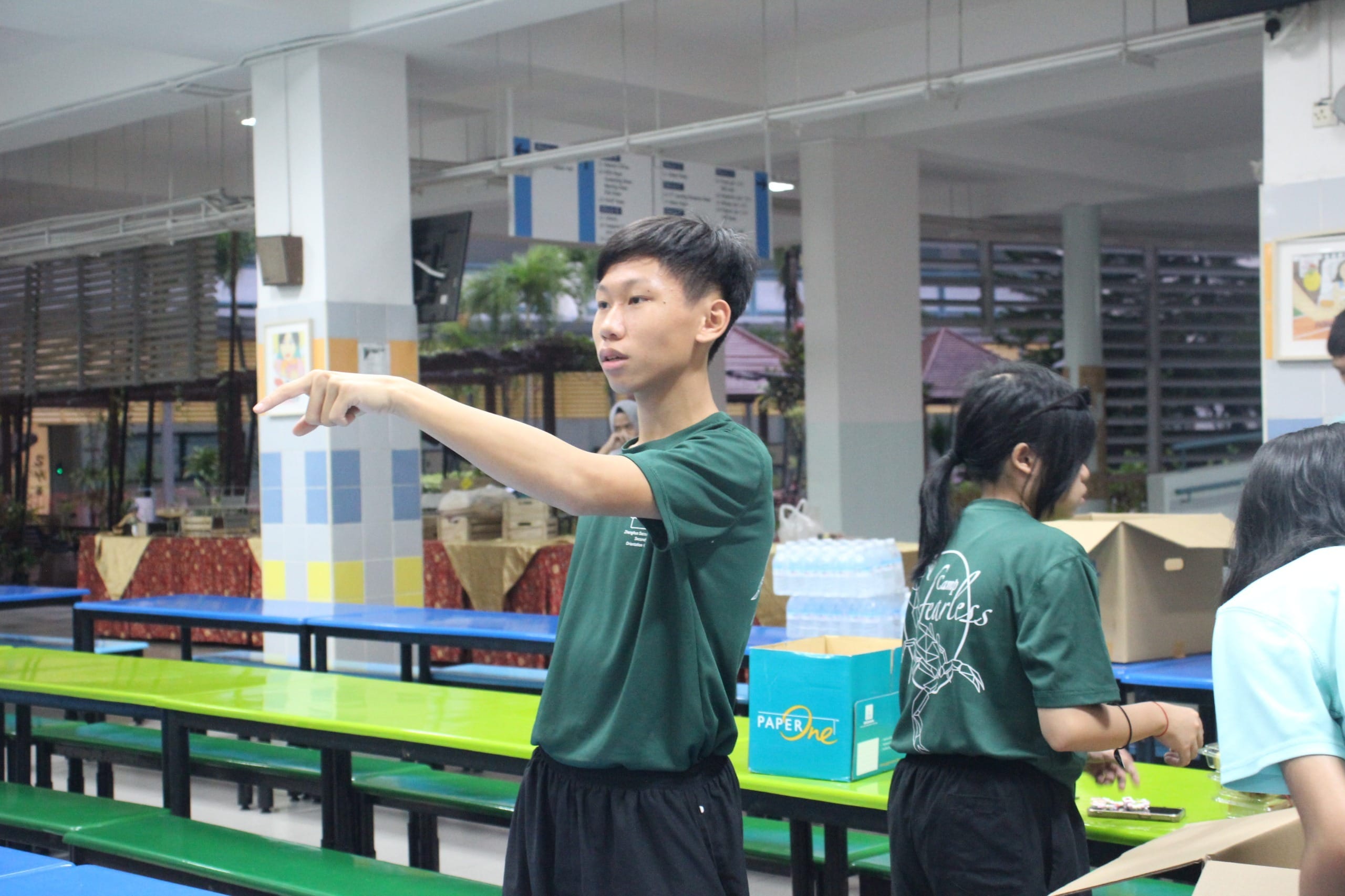 A teen in a green shirt points left in a school canteen with colorful benches and other students nearby.