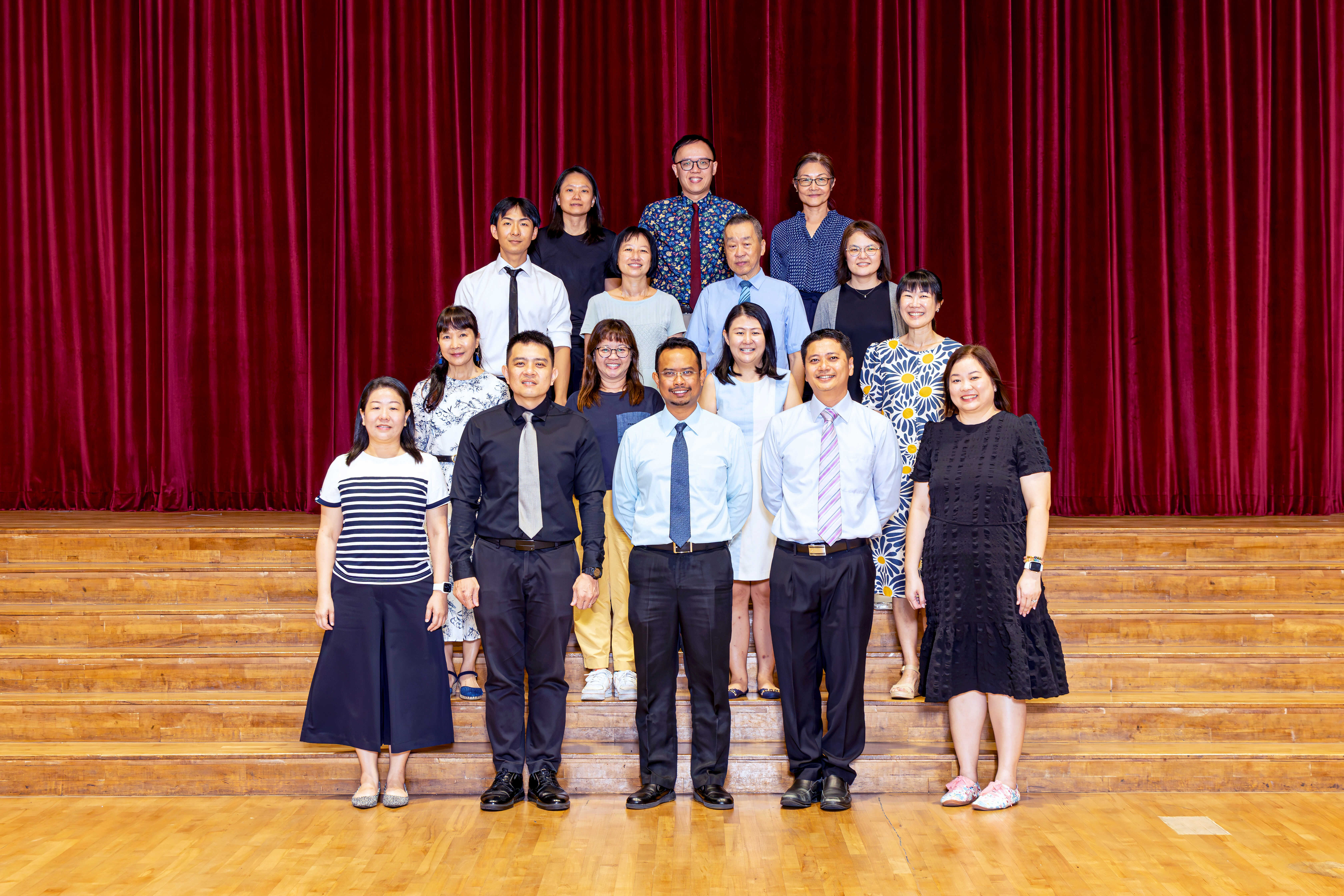 Group of about 15 adults posing in rows on wooden stage steps in front of a red curtain.