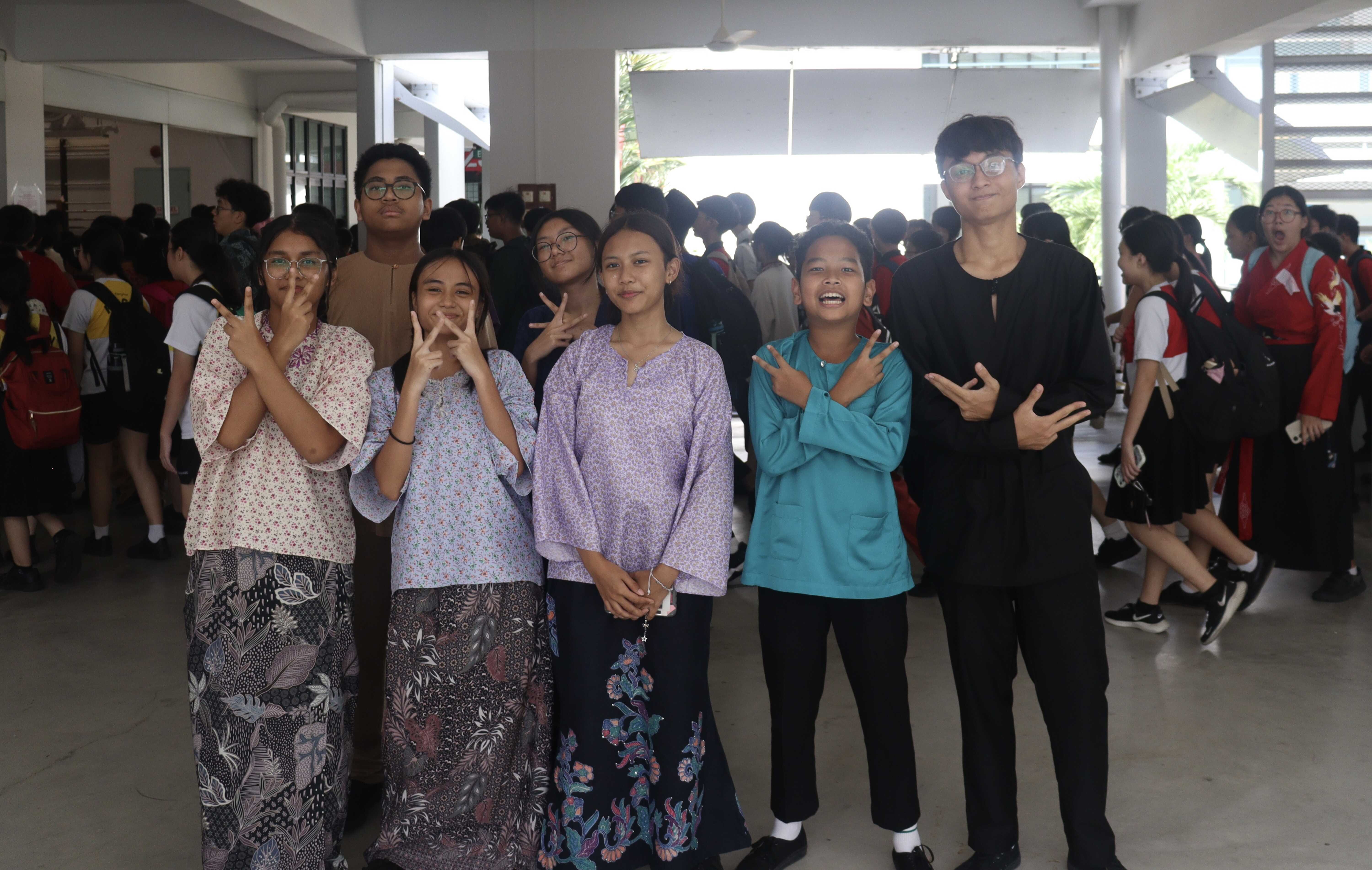 A group of students in traditional attire pose with peace signs in a school corridor with more students in the background.
