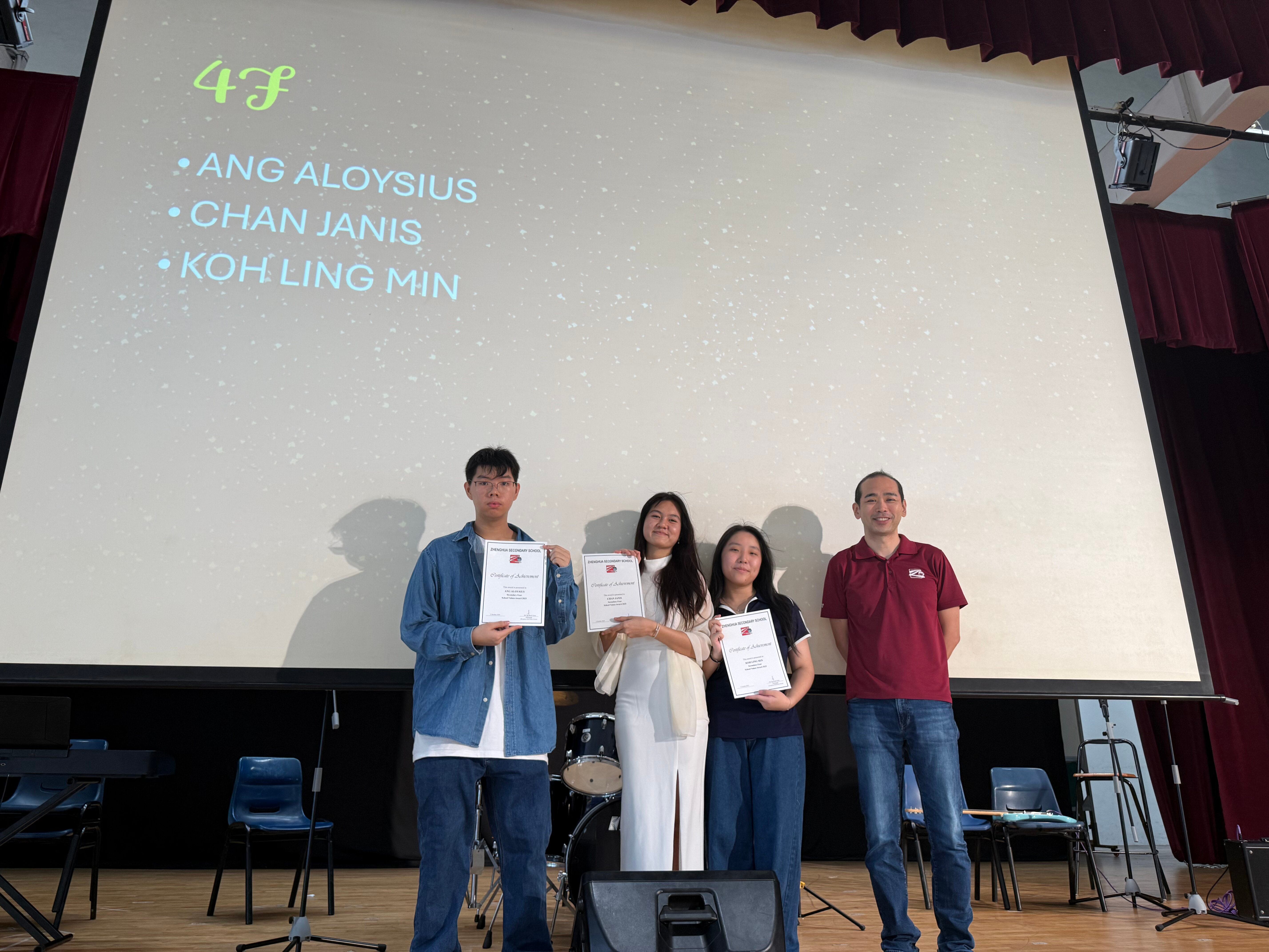 Four people stand on a stage holding certificates, with a screen behind showing names Ang Aloysius, Chan Janis, Koh Ling Min.