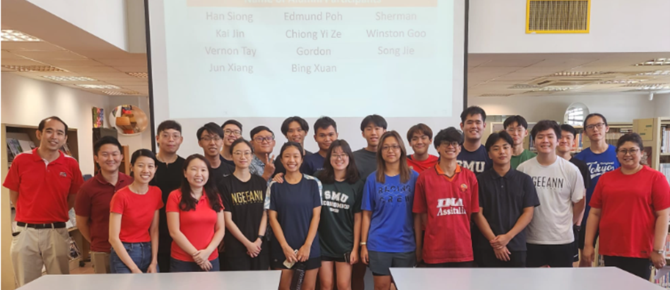 A large group of young people posing together indoors in front of a projection screen displaying student names.
