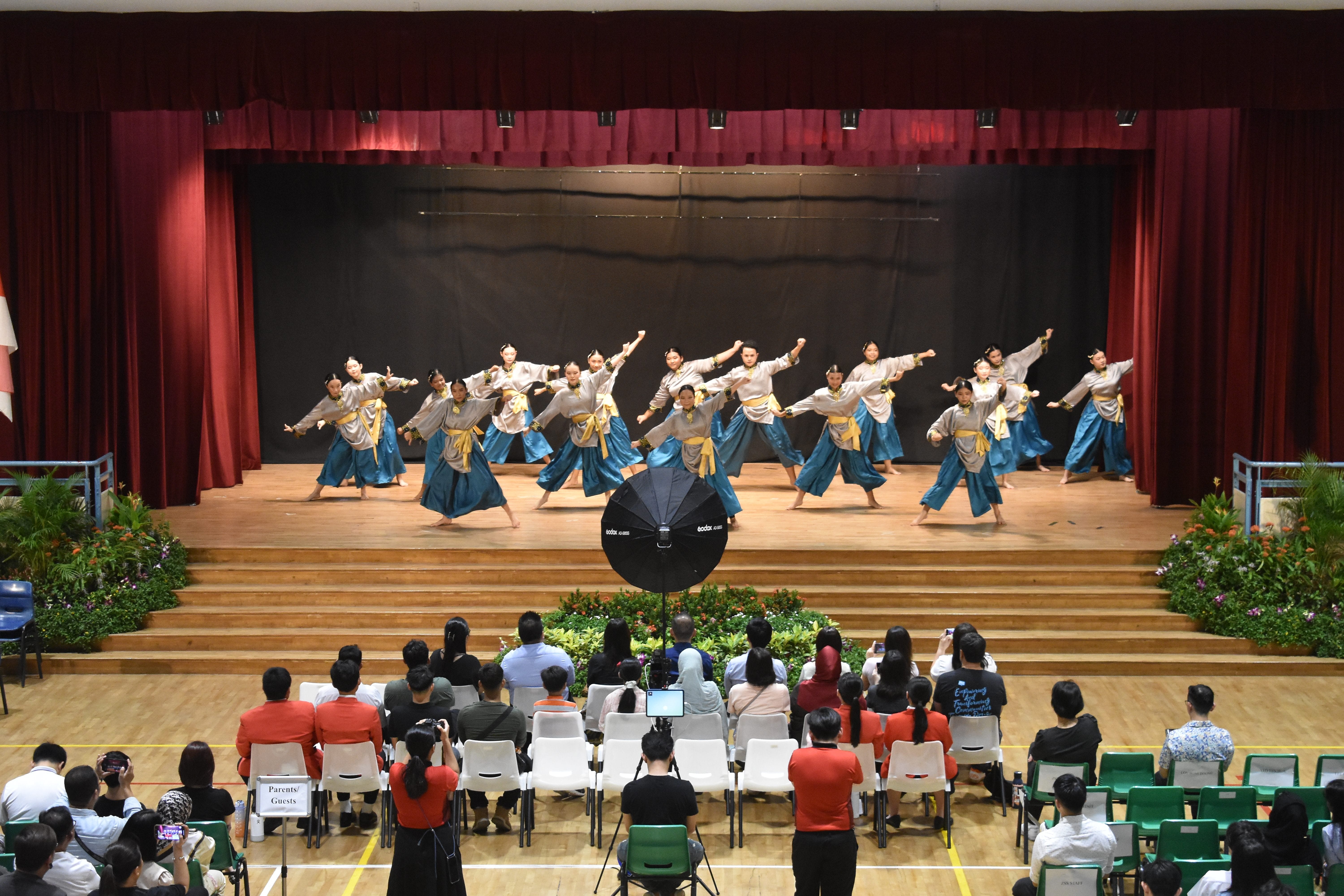 Group of dancers in blue skirts and yellow sashes perform on a stage before a seated audience in a hall.