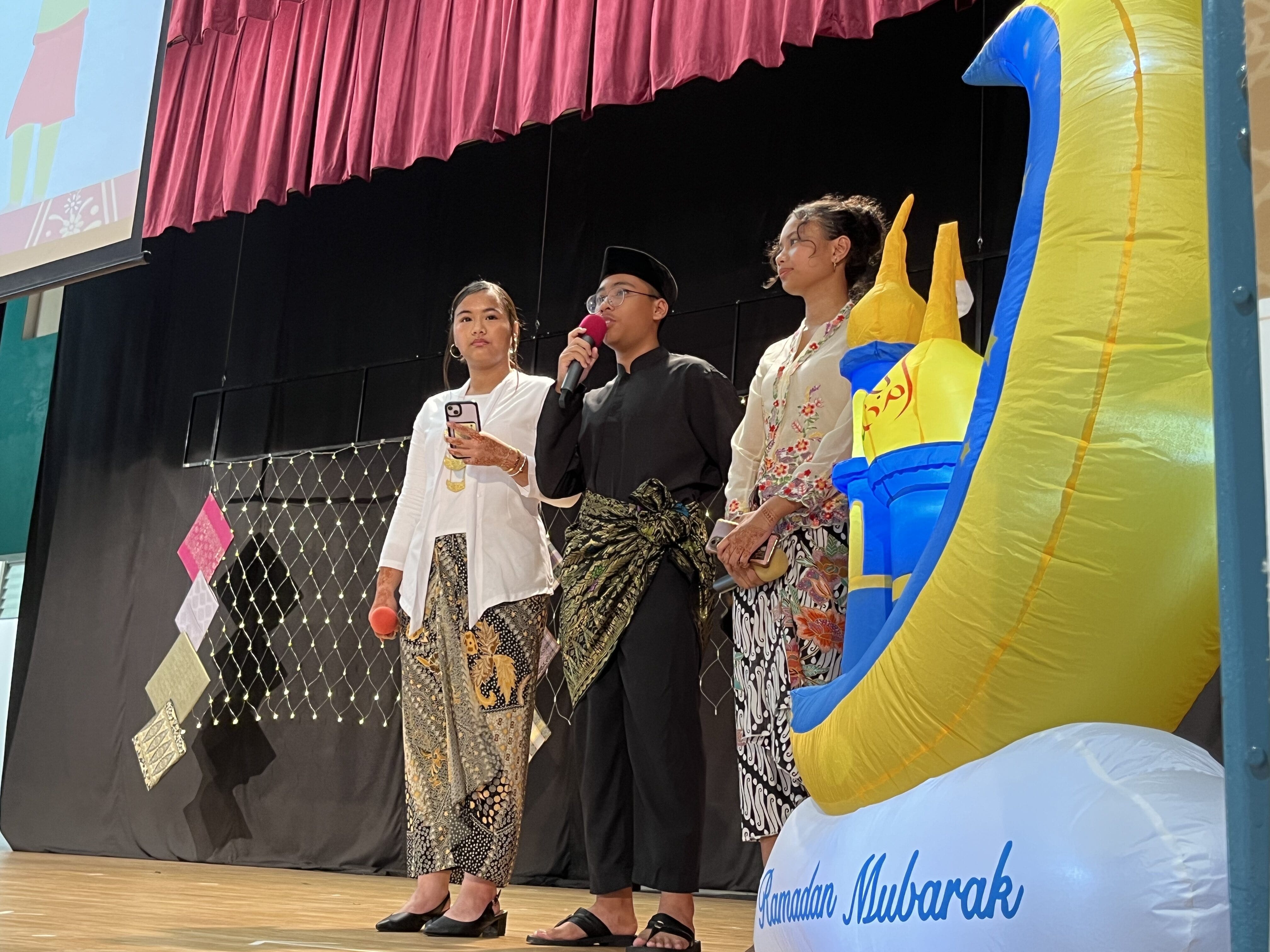 Three people in traditional attire stand on stage near a Ramadan Mubarak inflatable crescent moon decoration.
