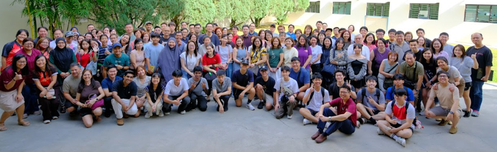 Large group of people posing together outdoors near trees and a building for a group photo.