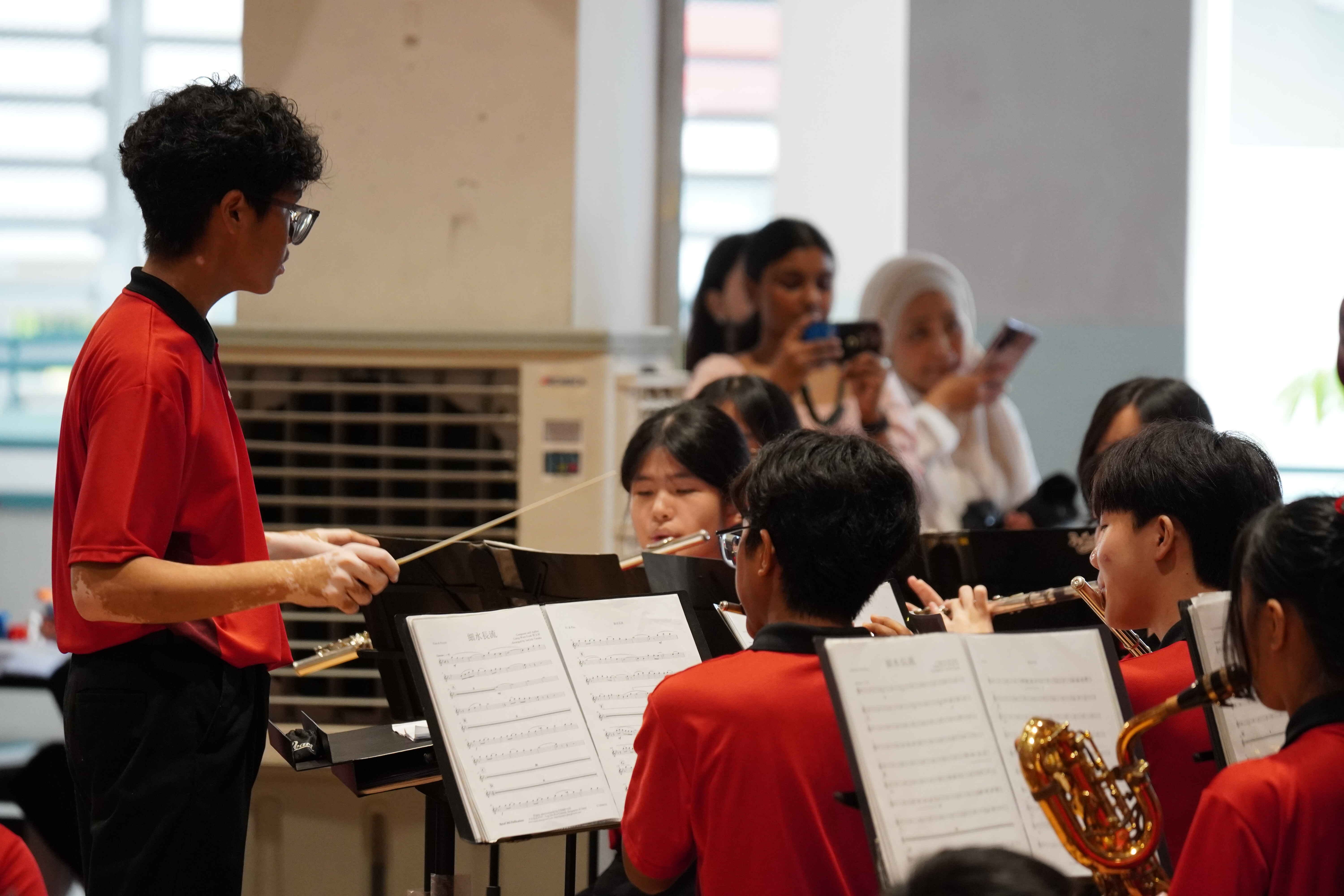 A young conductor in a red shirt leads student musicians with flutes and saxophone reading sheet music indoors.