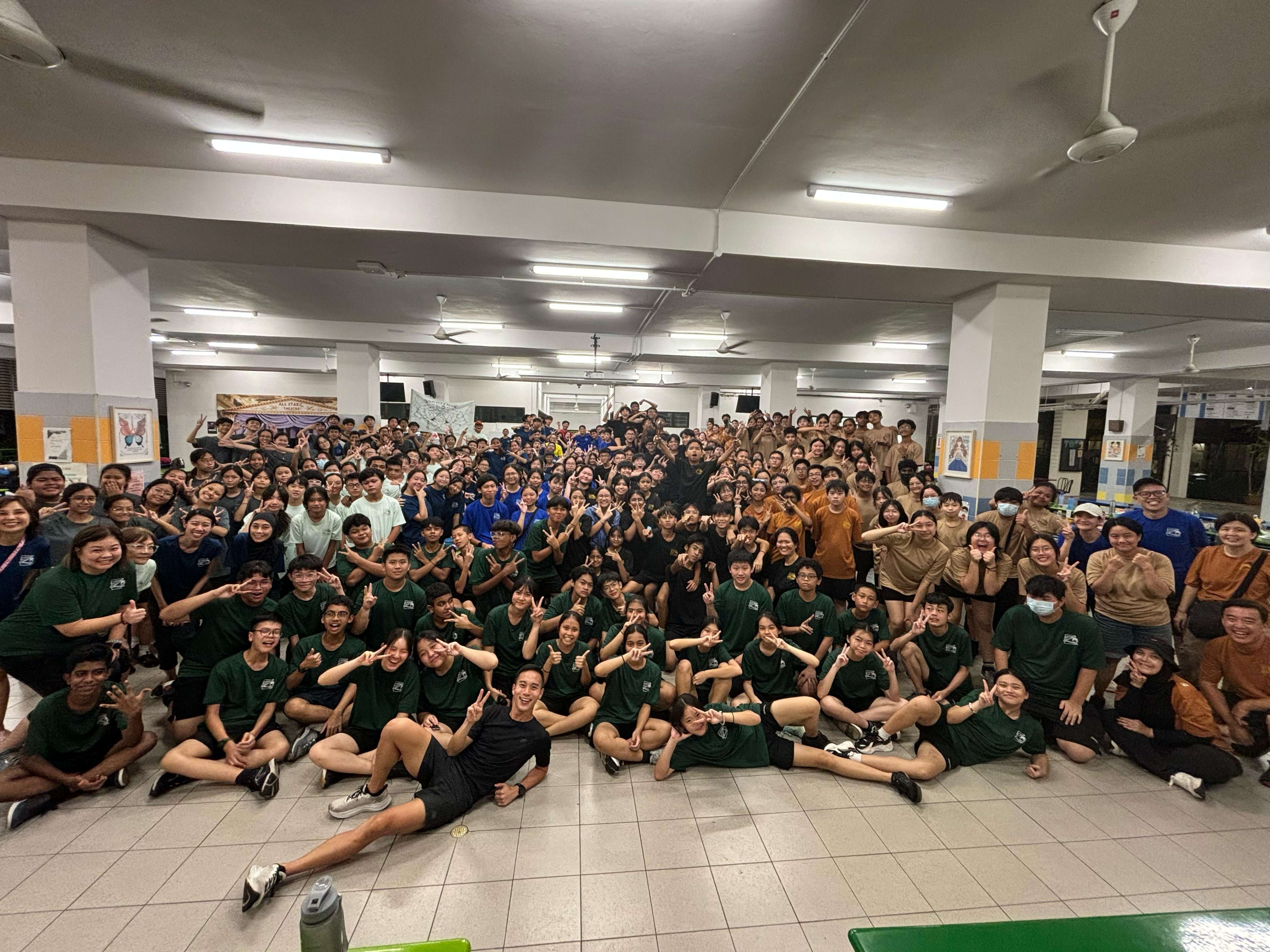 Large group of students in matching colored shirts posing together indoors, many making peace signs.