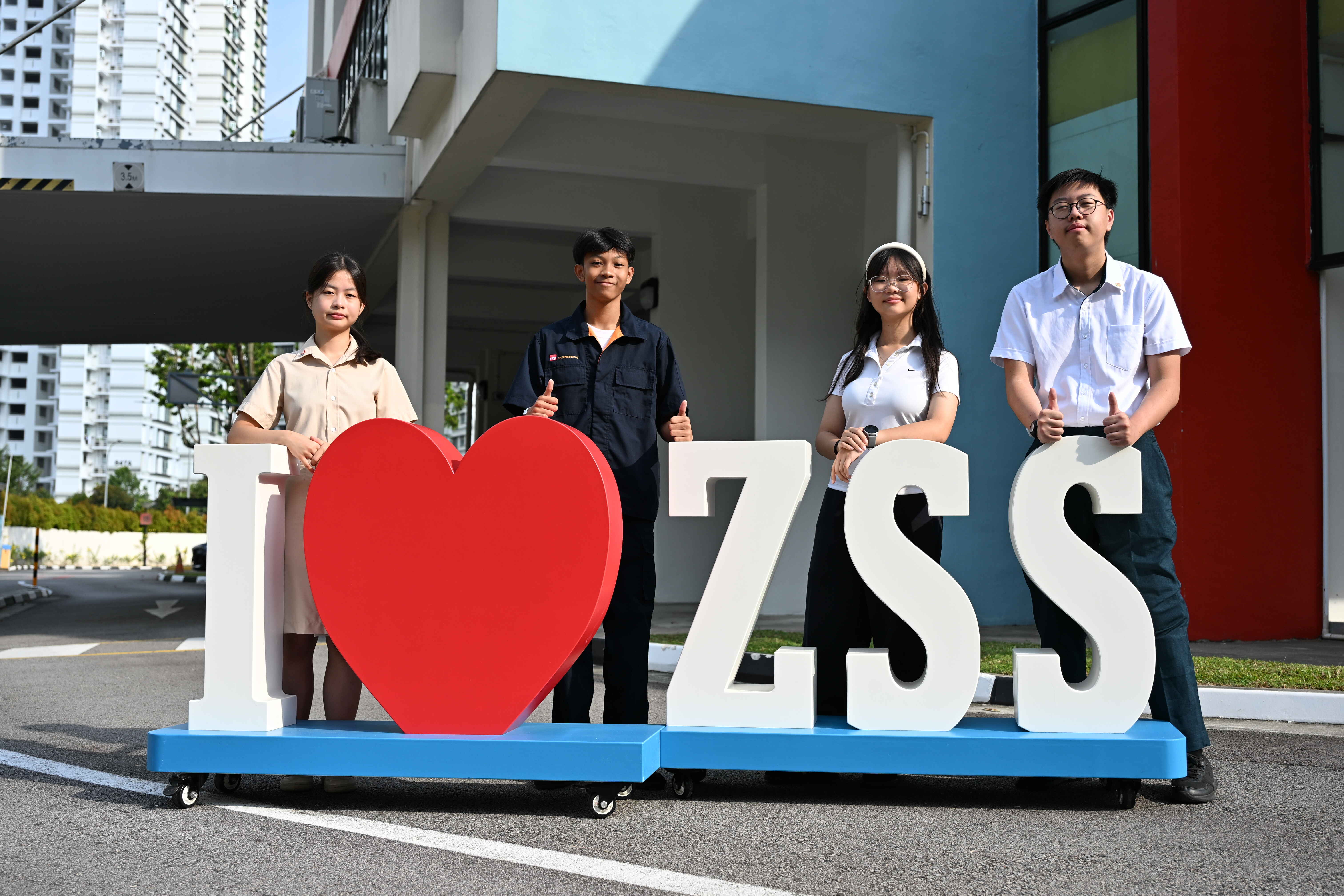 Four students posing behind a large I Love ZSS sign with a red heart outdoors near a school building.
