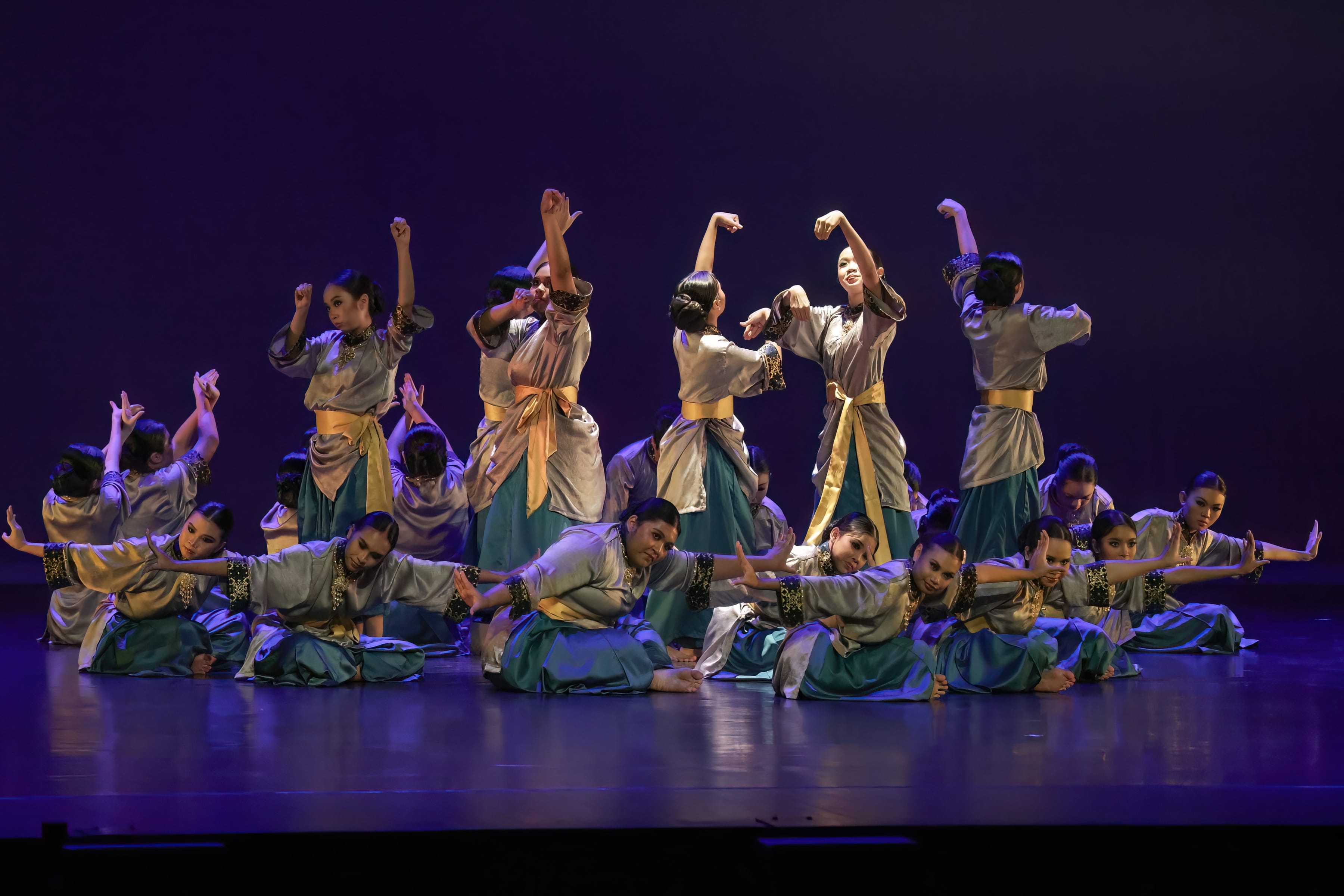 A large group of dancers in silver and teal costumes with gold sashes perform on a purple-lit stage.