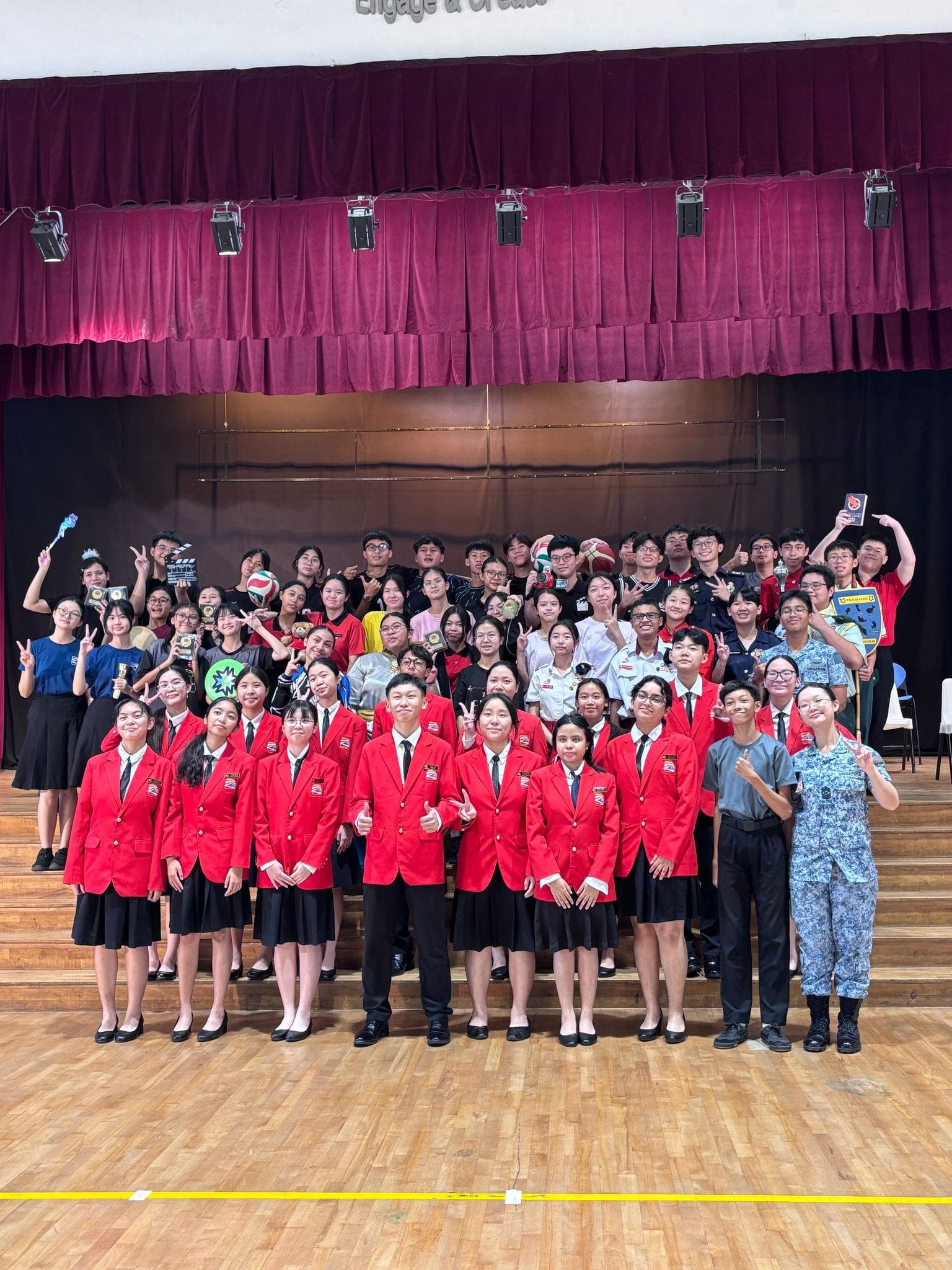 Large group of students in red blazers and various uniforms posing on a school hall stage with trophies.