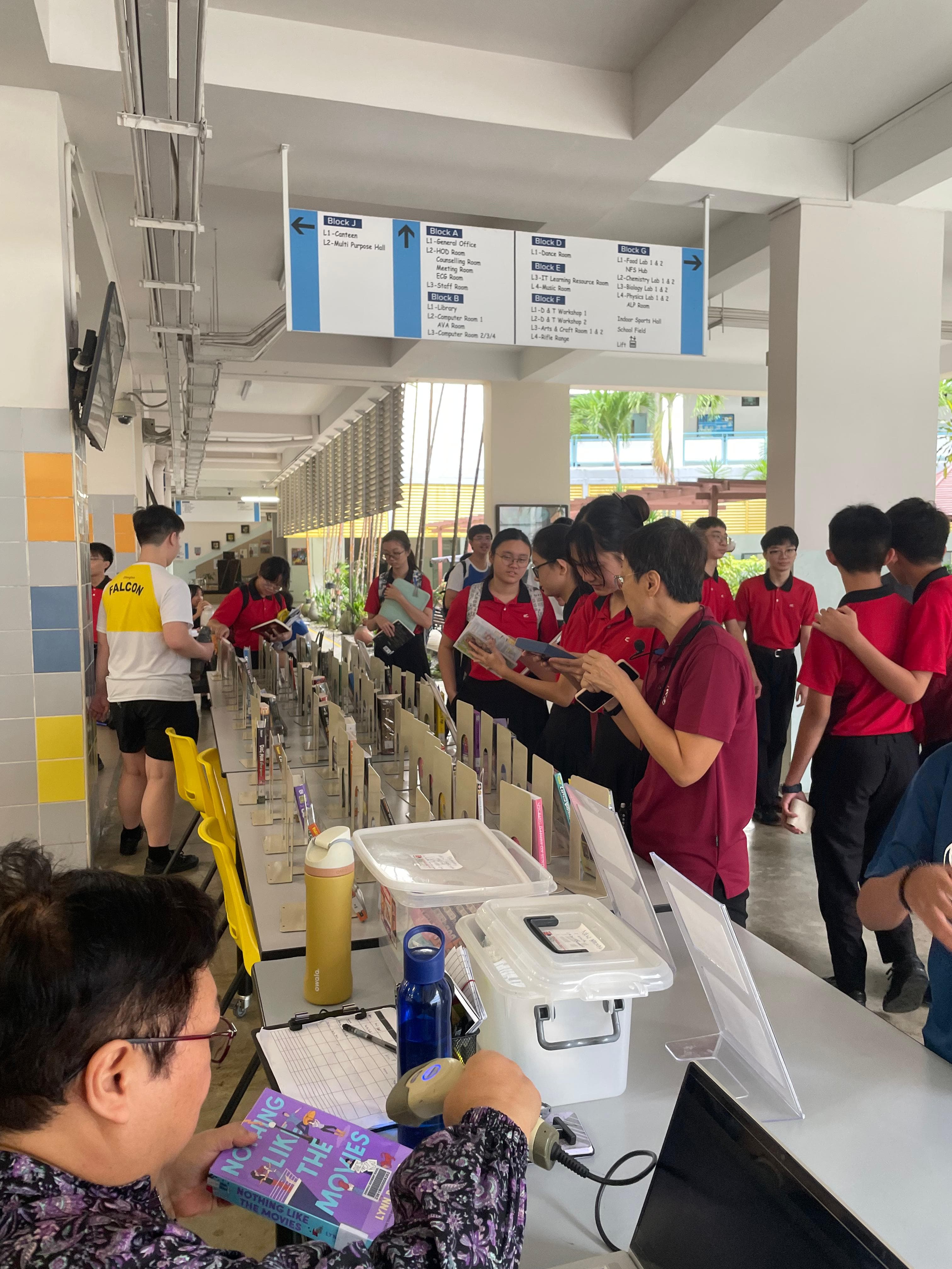 Students in red uniforms browse books at a school corridor counter with directional signs overhead.