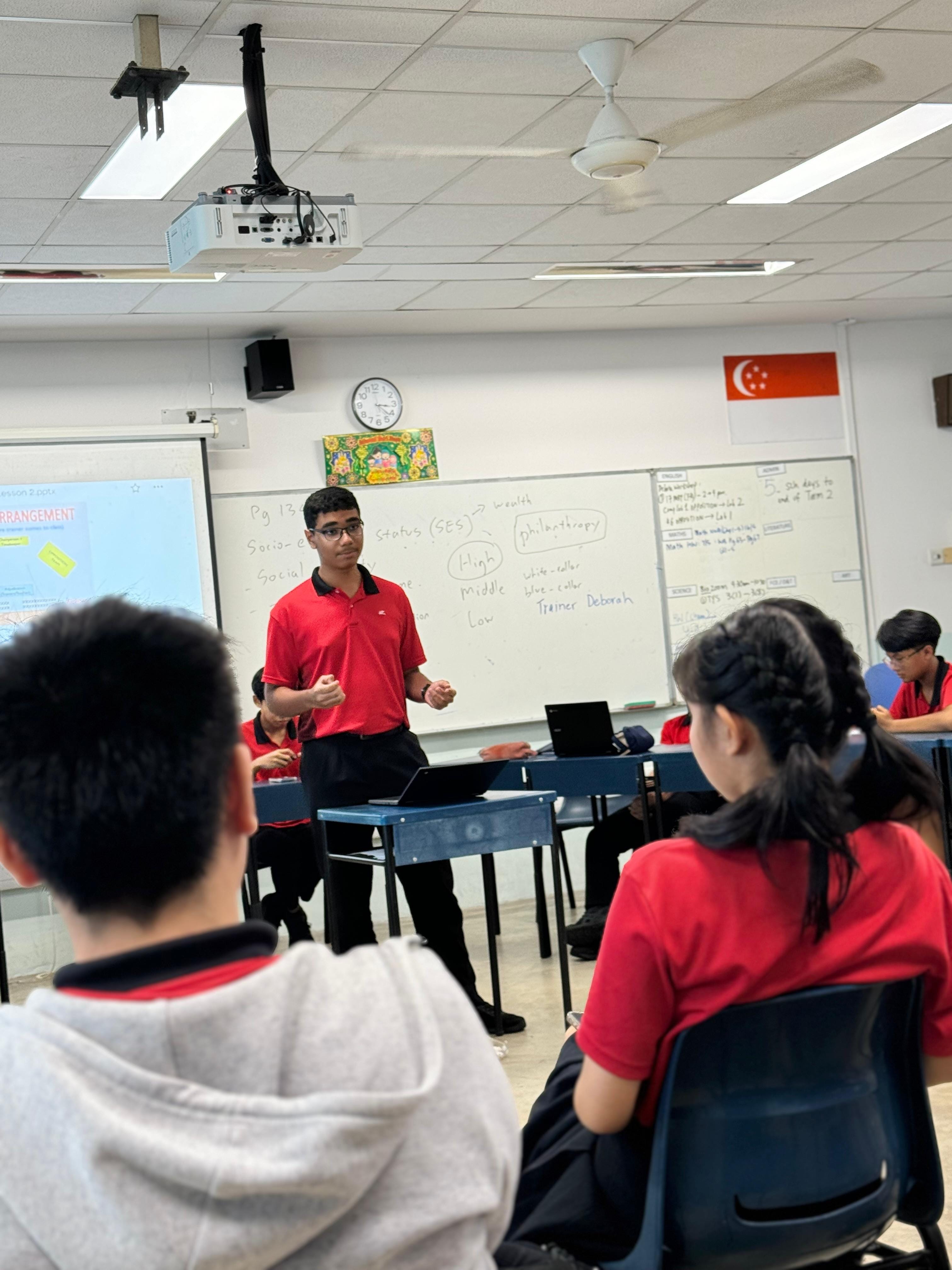 A student in a red uniform presents at a whiteboard in a classroom with a Singapore flag on the wall.