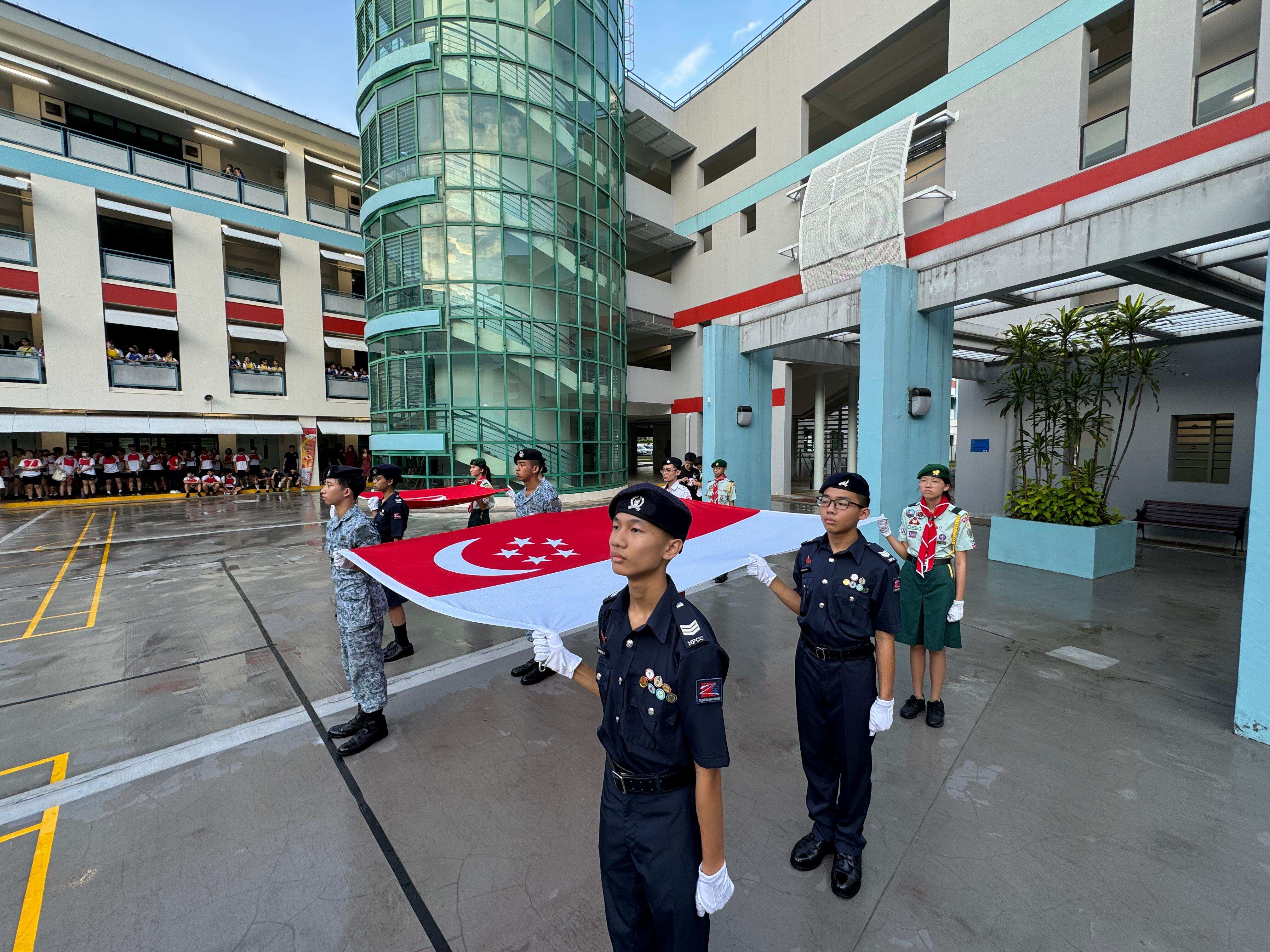 Students in uniform carry the Singapore flag during a ceremony in a school courtyard with spectators watching.