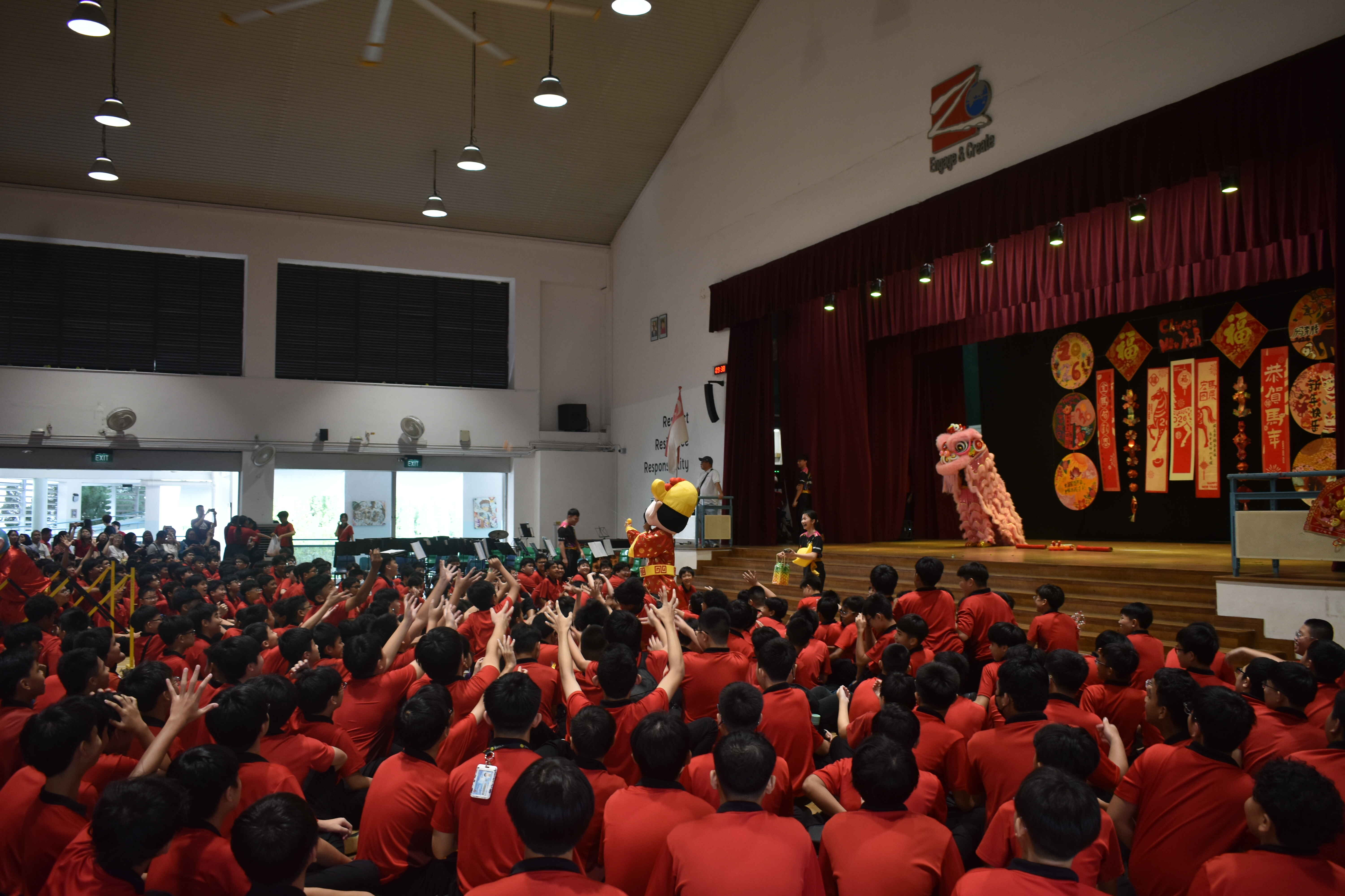 Students in red shirts watch a lion dance performance on a decorated stage during a Chinese New Year school event.