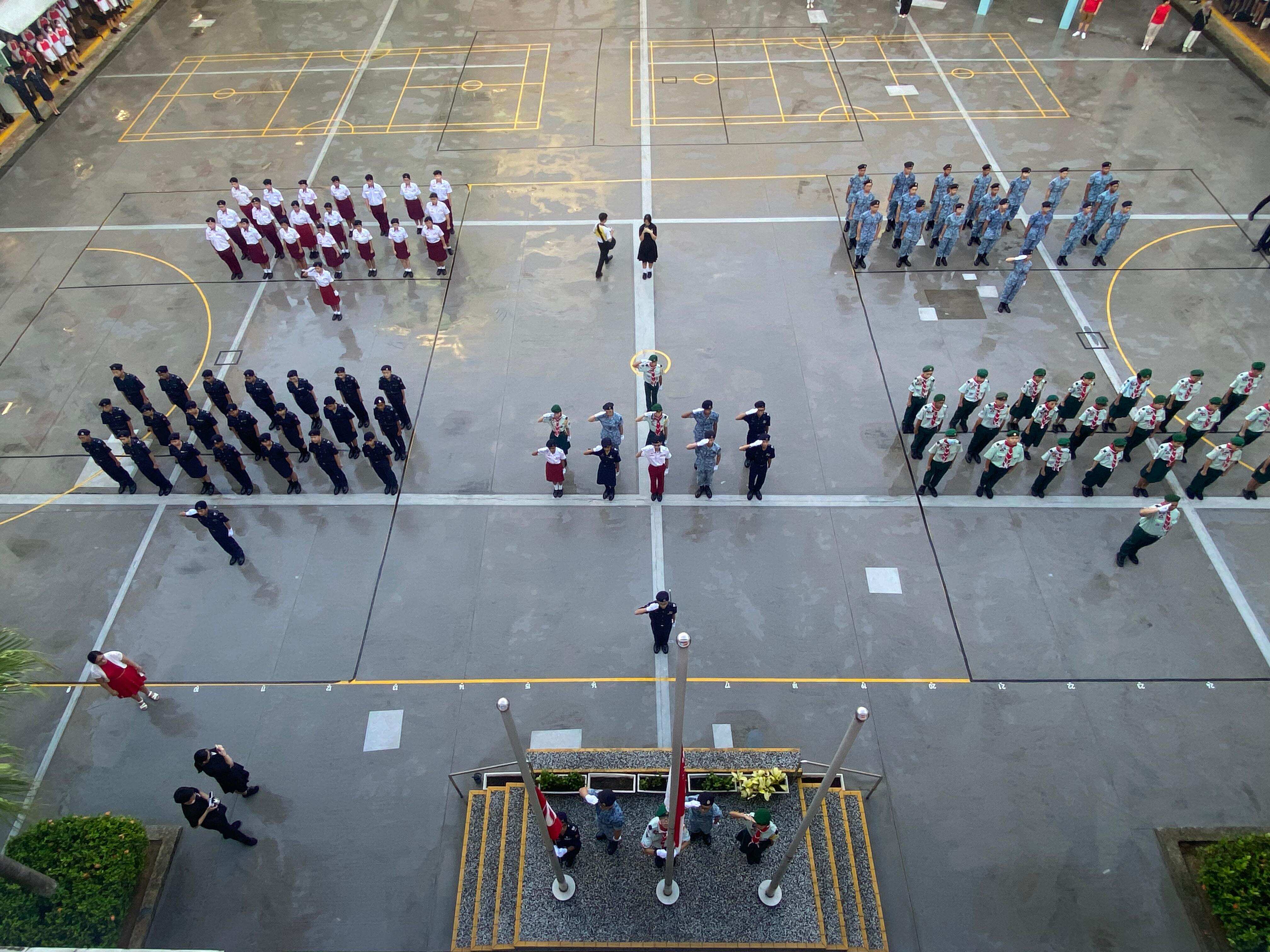 Aerial view of a school parade with uniformed student groups standing in formation on a courtyard near flag poles.