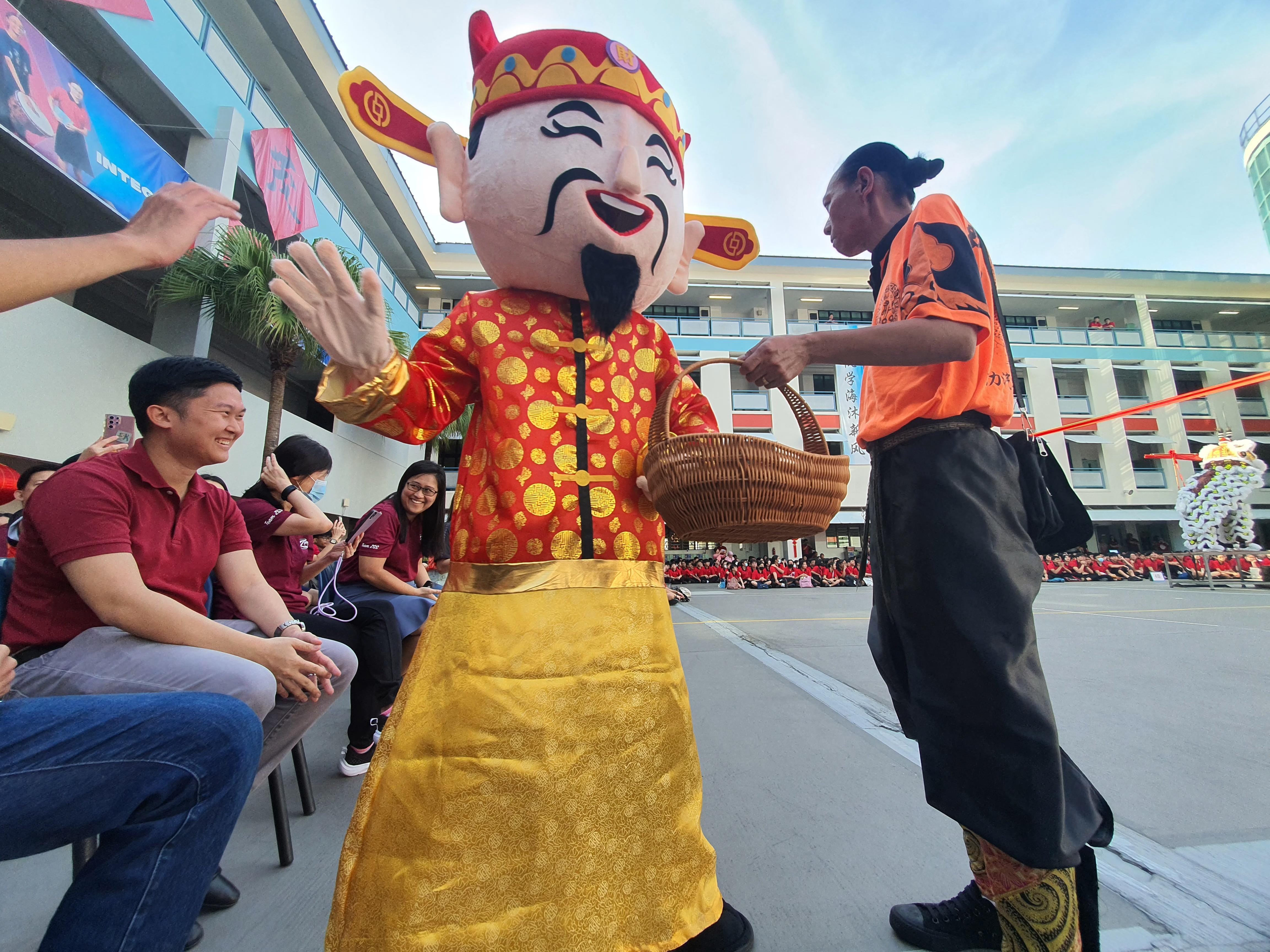 A large Chinese God of Wealth mascot in red and gold interacts with seated spectators at an outdoor school event.