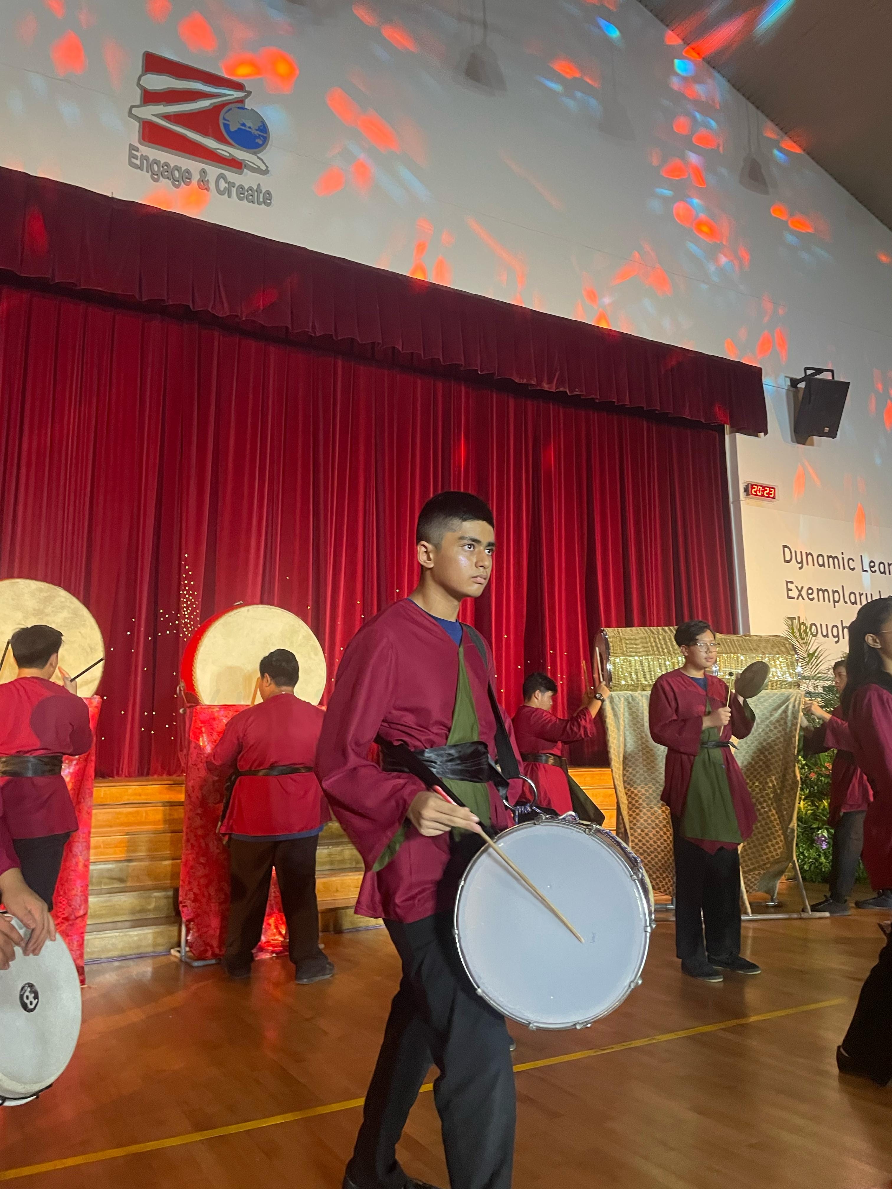 Young performers in red and green costumes play drums on a stage with red curtains and colorful light projections.