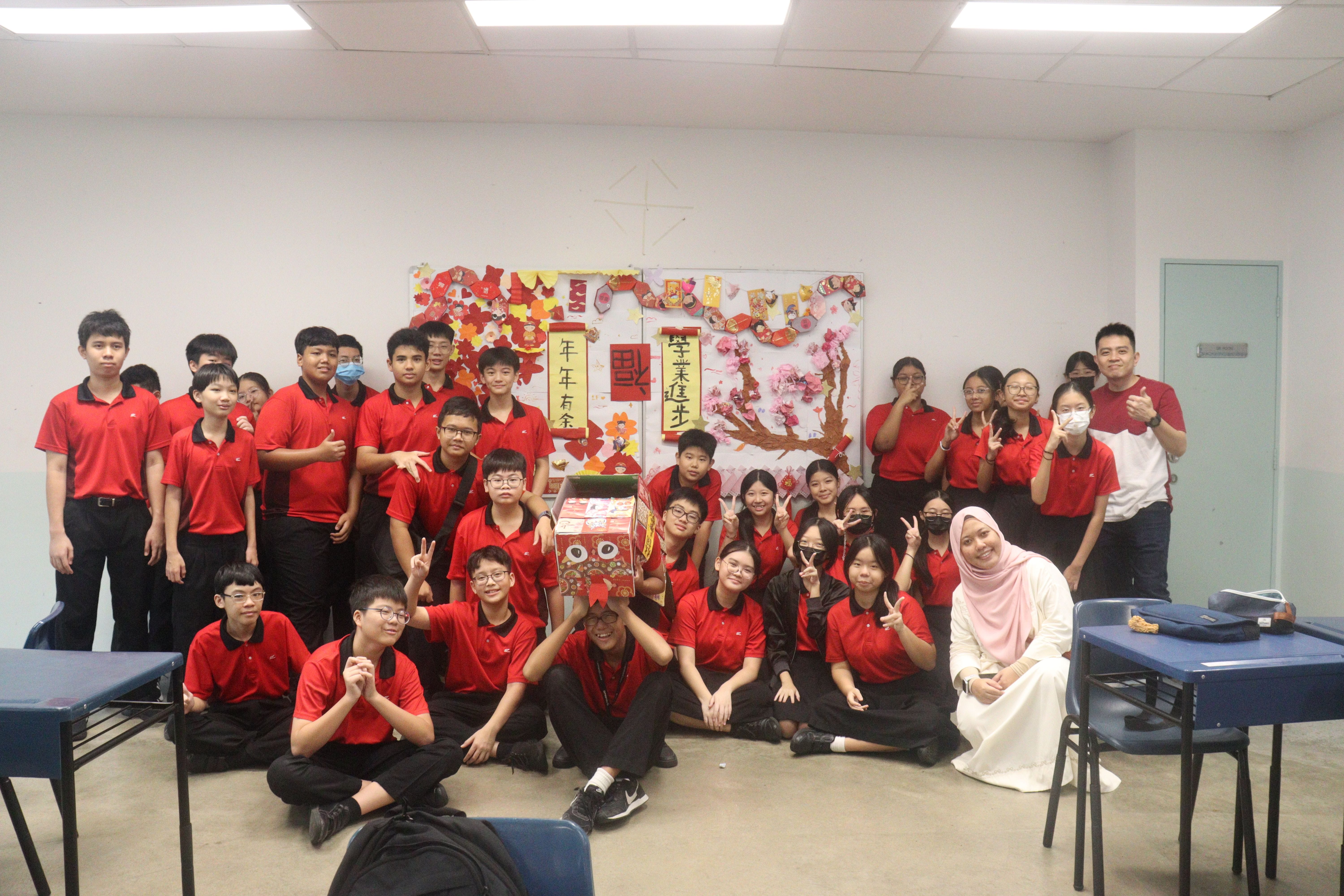 A large group of students in red uniforms pose in a classroom decorated with Chinese New Year decorations.