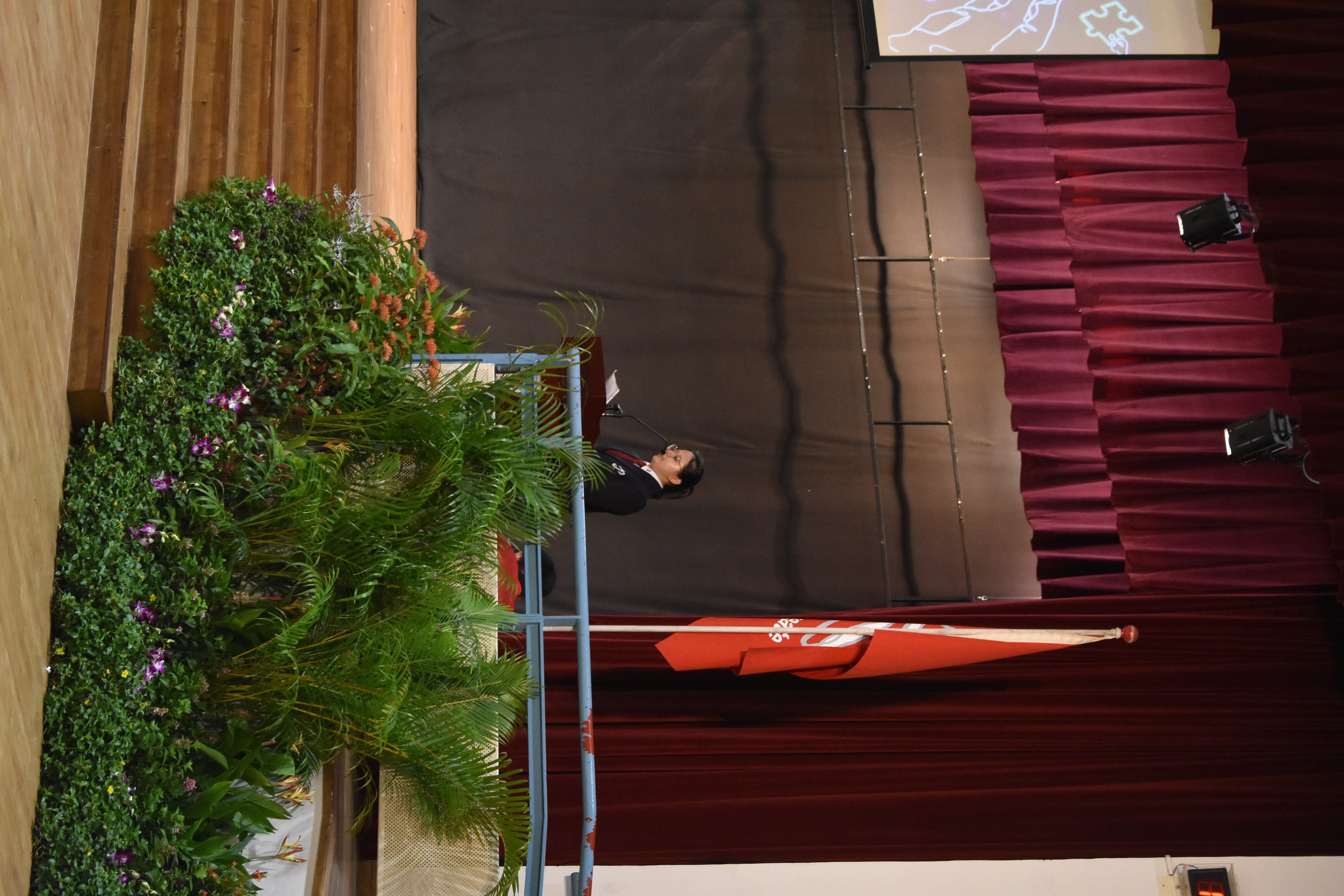 A person in a dark blazer speaks at a podium on a stage with red curtains, an orange flag, and plants below.