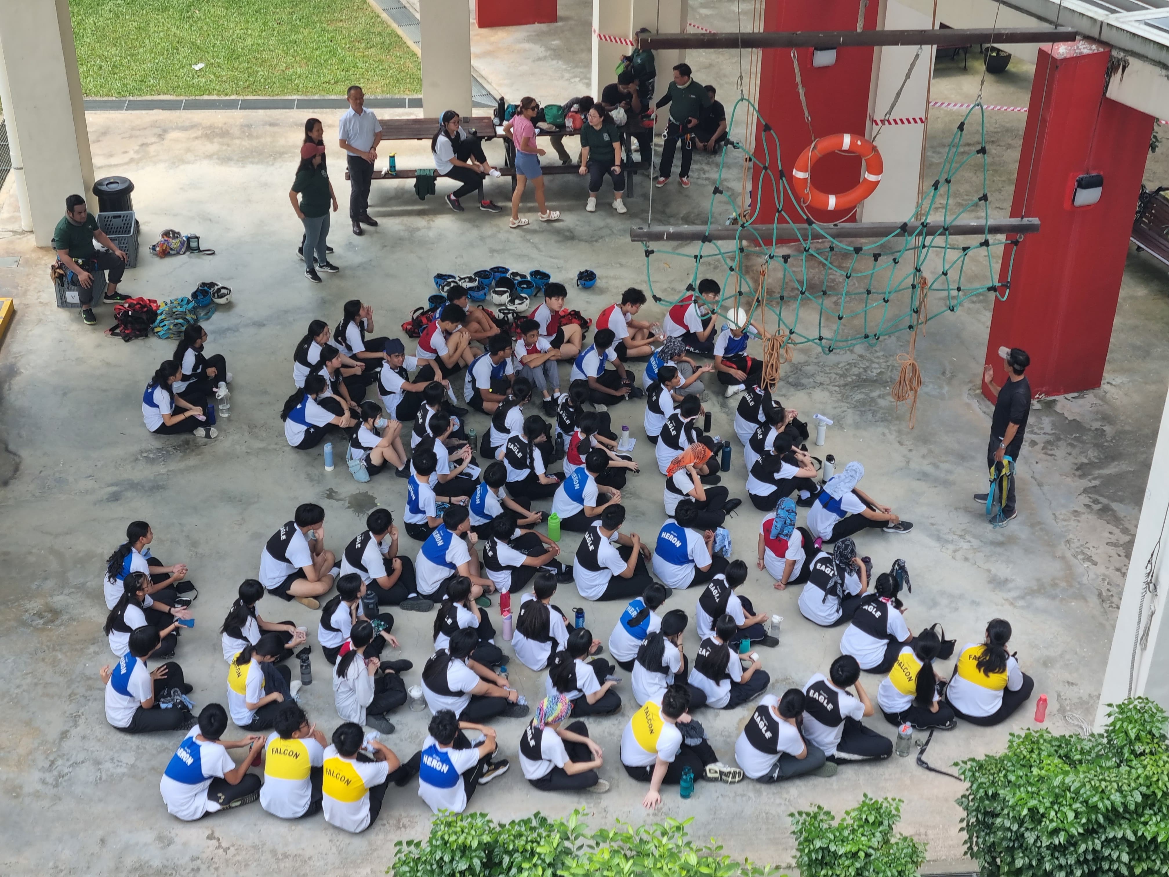 Large group of students in white and black uniforms sitting on a courtyard floor near a rope climbing structure.