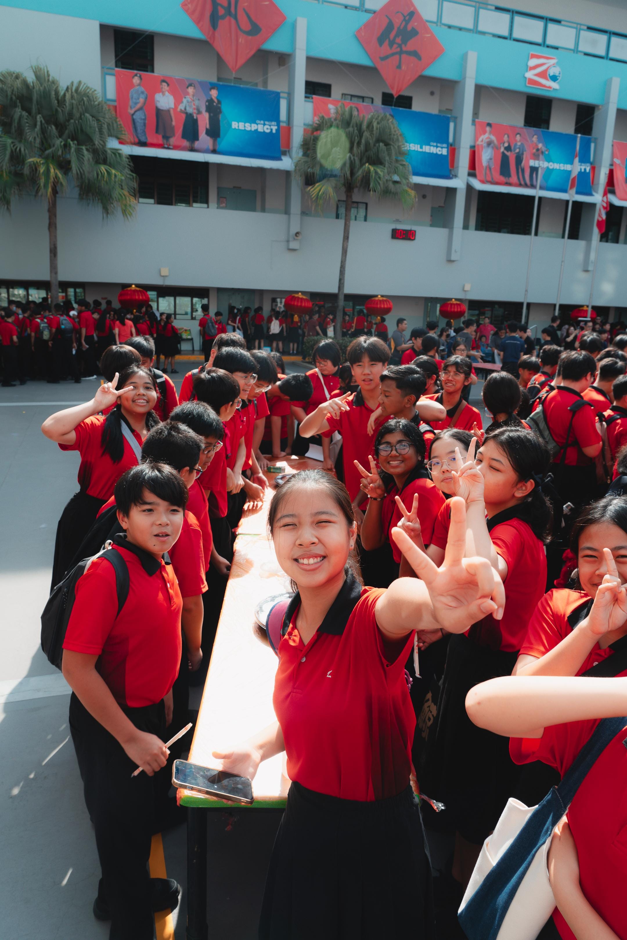 A large group of students in red and black uniforms smile and flash peace signs in a school courtyard.