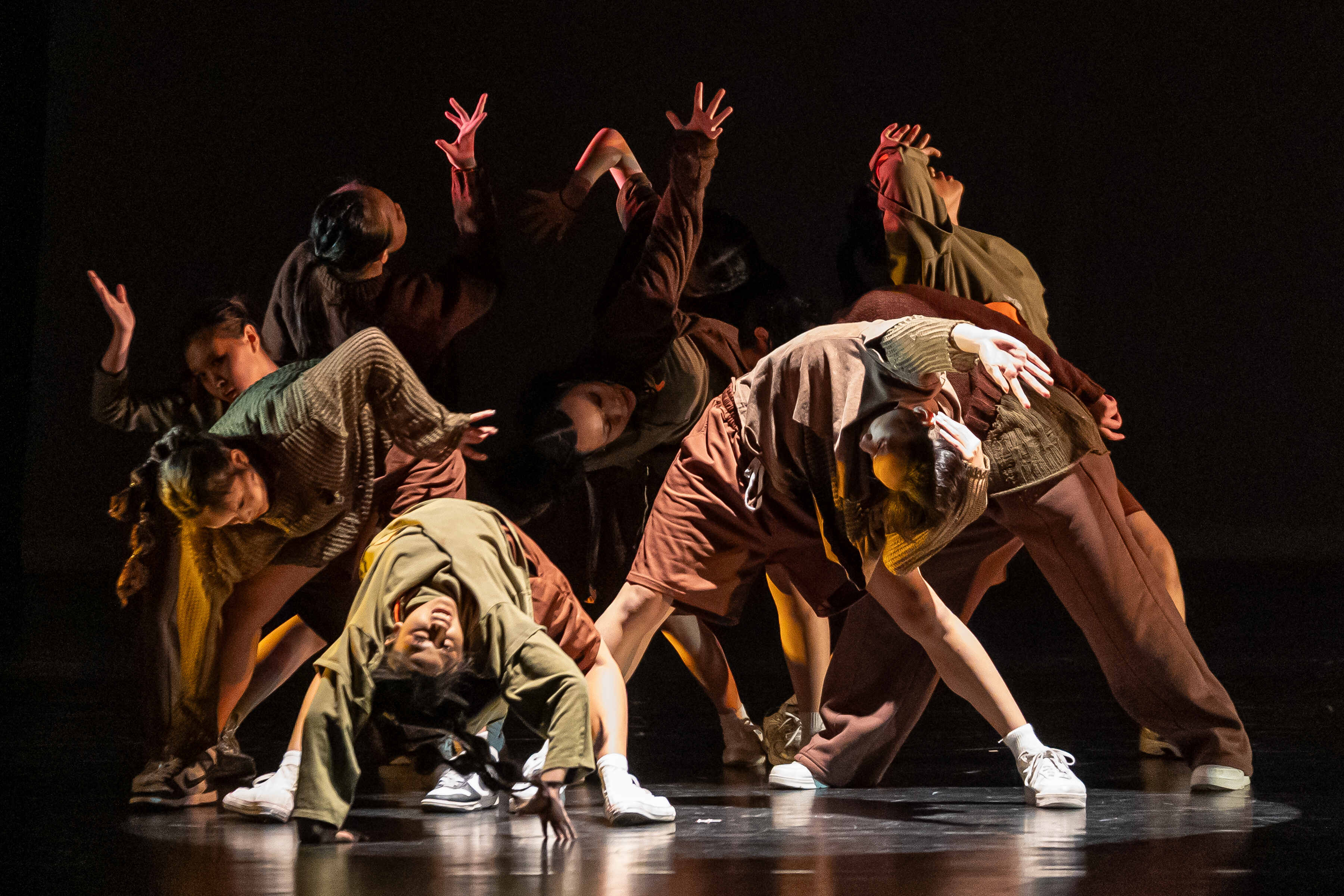Group of dancers in earth-toned clothing performing dramatic moves on a dark stage, one dancer bent backward near the floor.