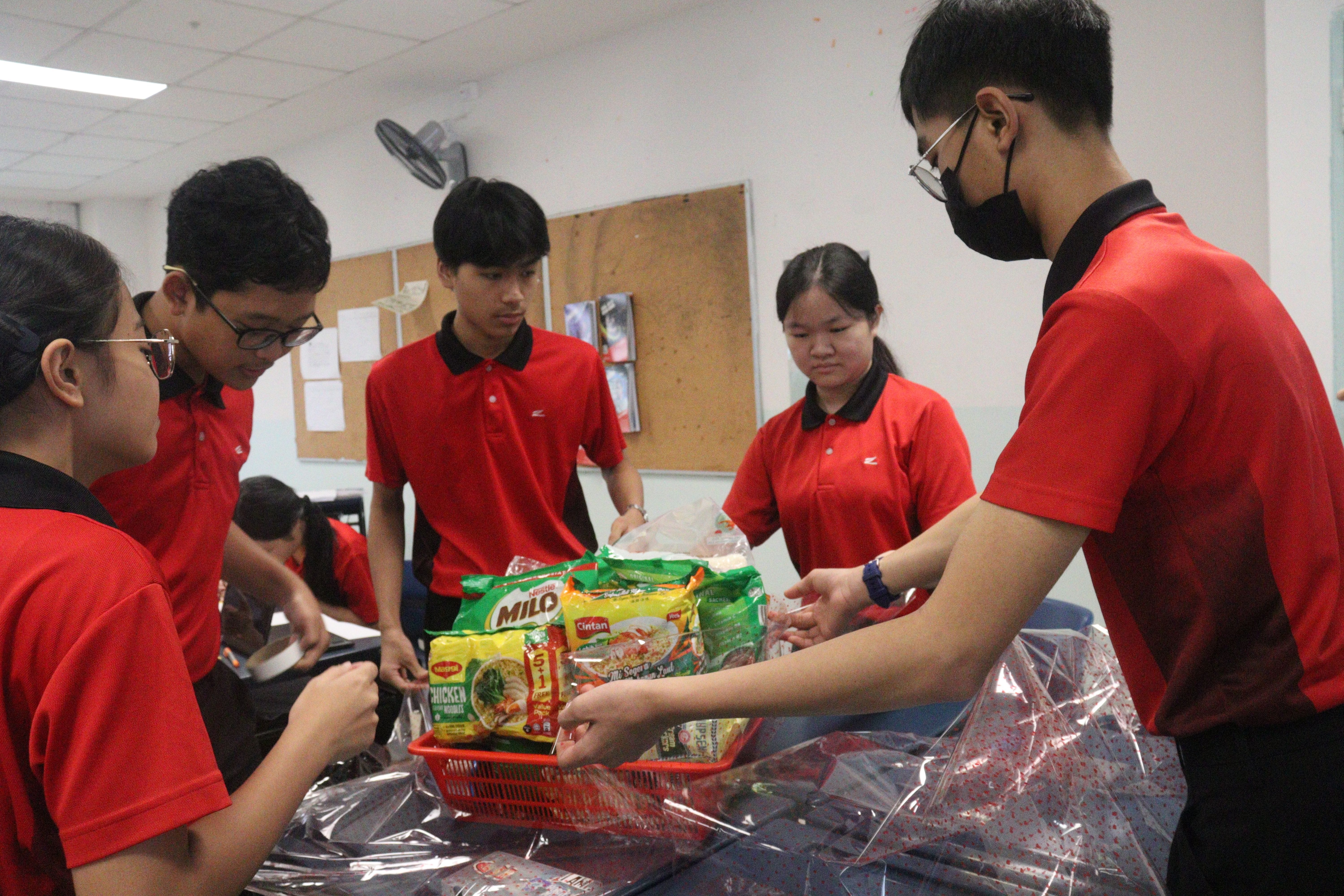 Students in red uniforms packing food items including Milo and instant noodles into plastic bags at a table.