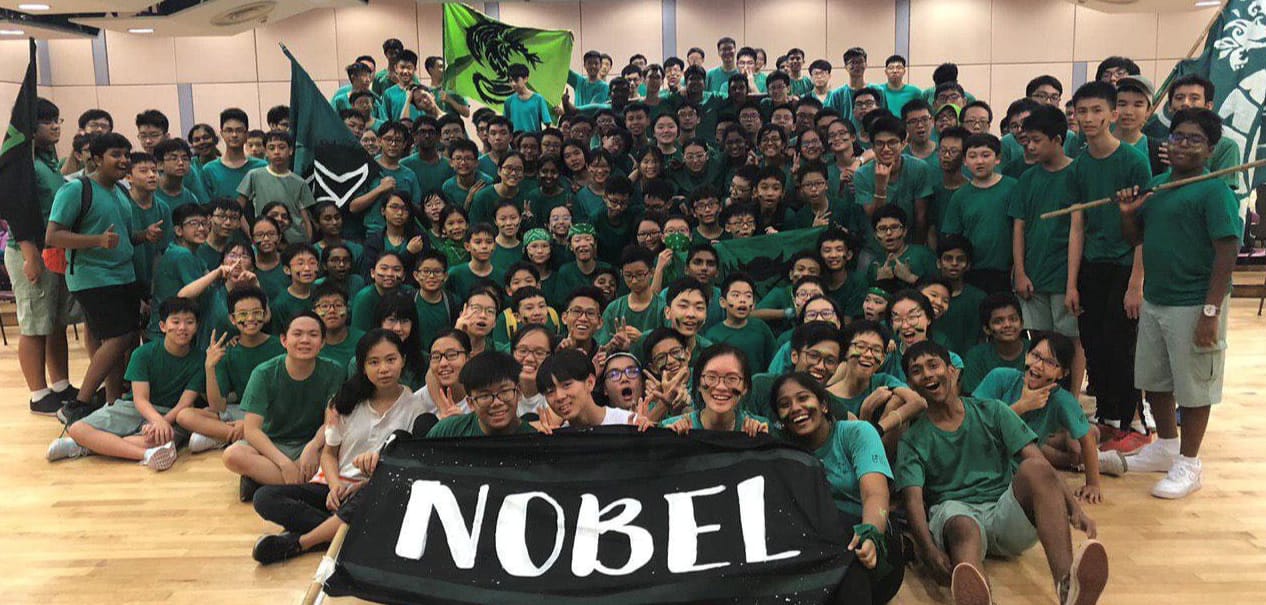 Large group of students in green shirts posing together indoors, holding a black banner reading NOBEL and green flags.
