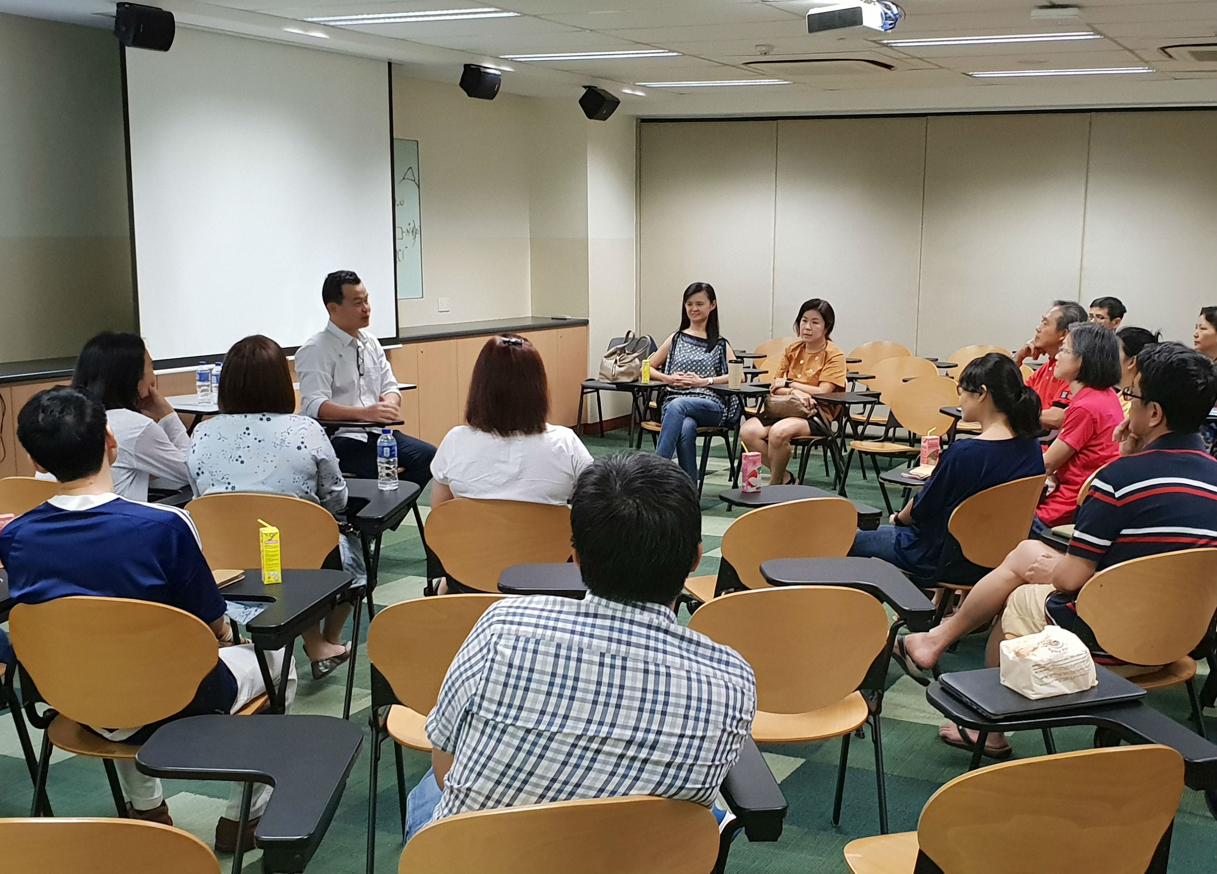 A group of adults sit in chairs arranged in a circle in a classroom with a projector screen nearby.