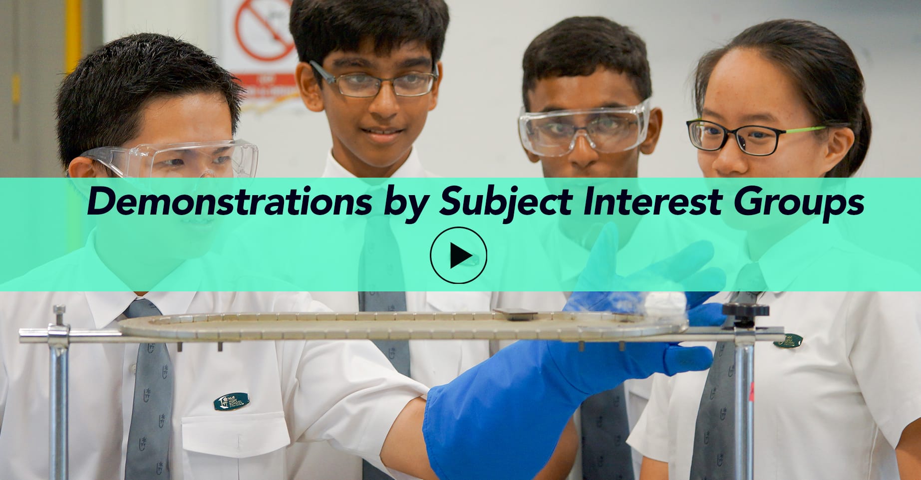 Four students in lab coats and safety goggles watch a science demonstration with a rack and blue gloves.