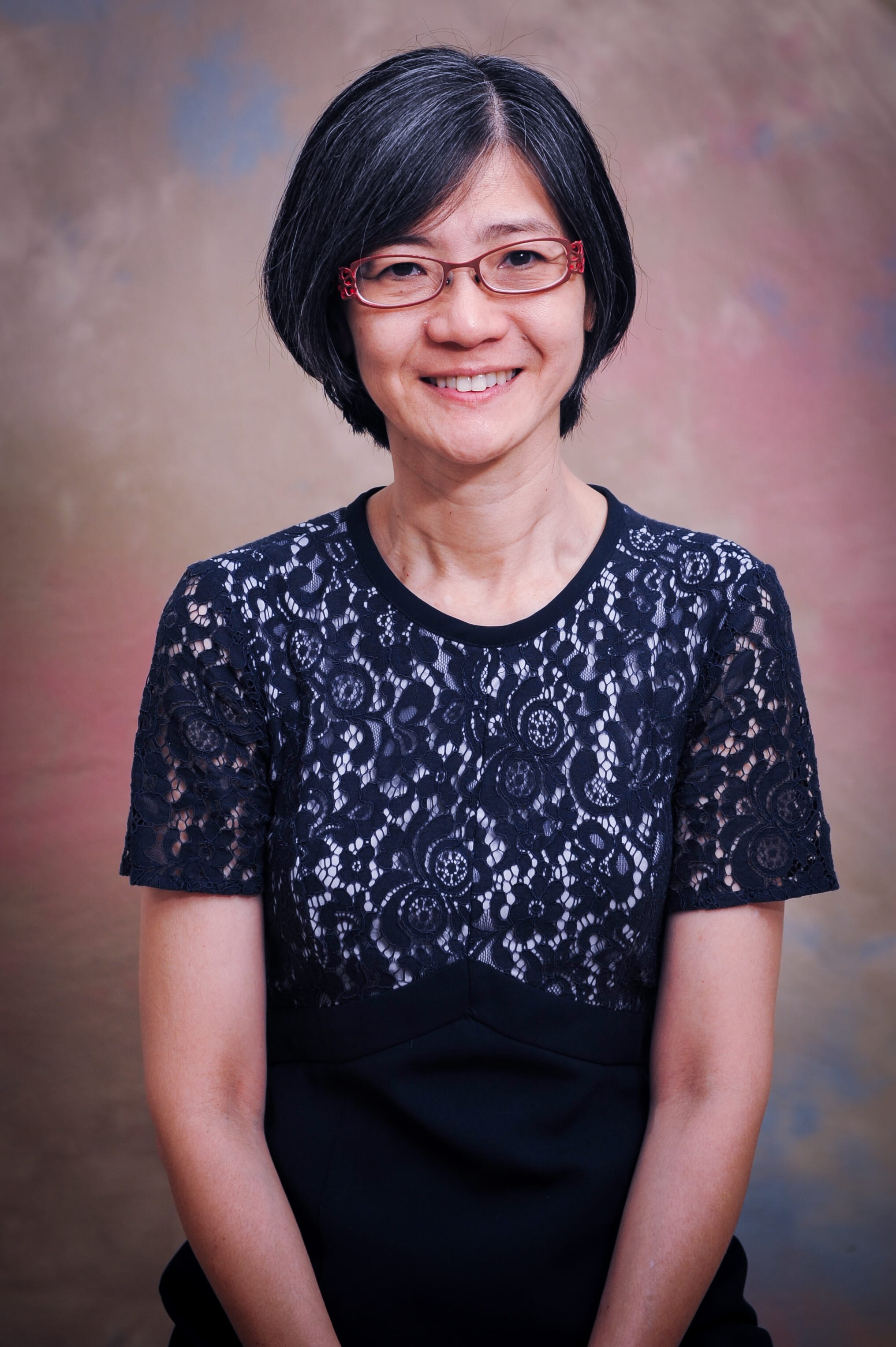 A smiling woman with short black hair and red glasses wearing a black lace top, posed against a muted background.