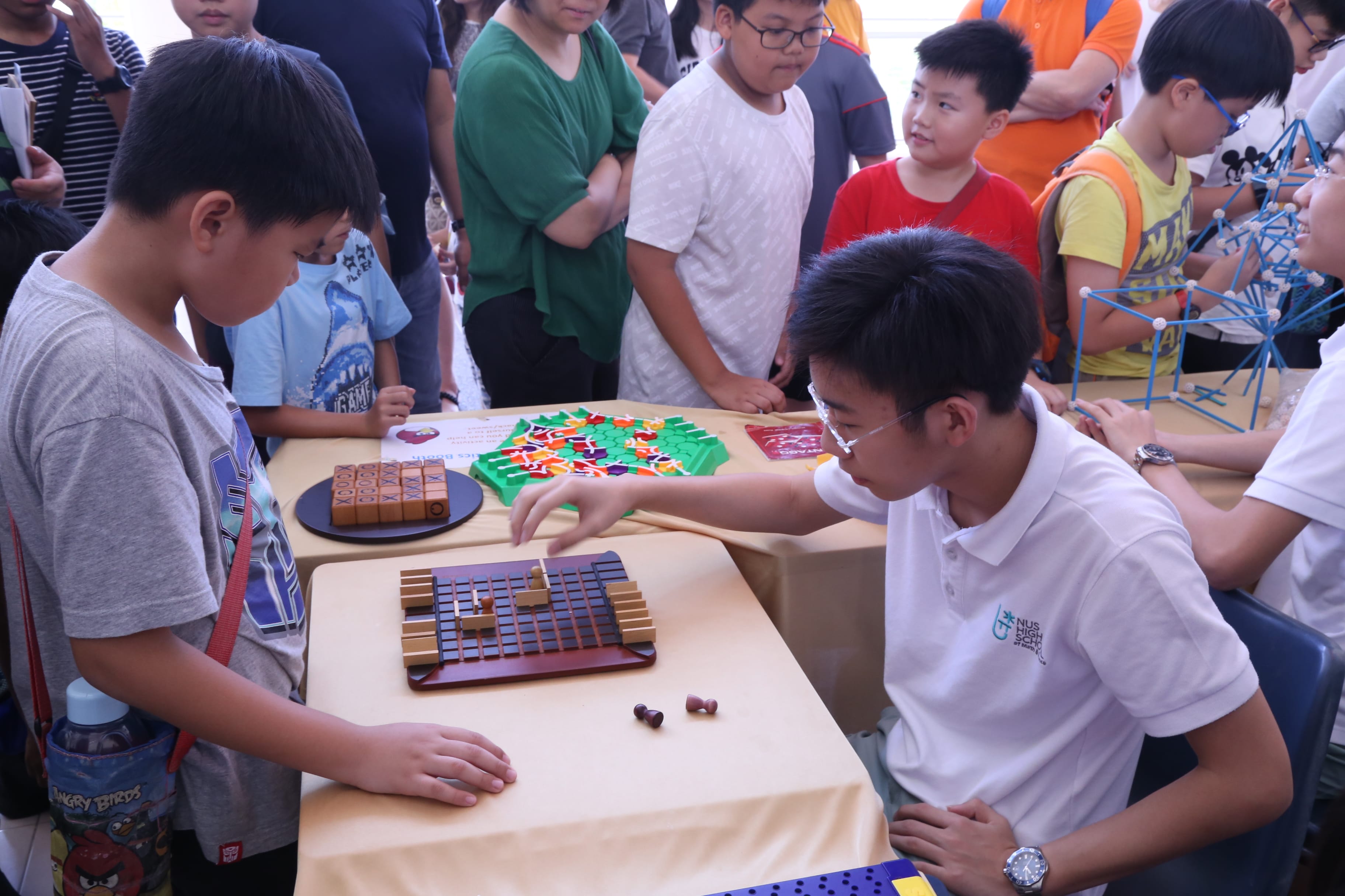 Boy in glasses moves pieces on a tabletop board game while children gather around watching.