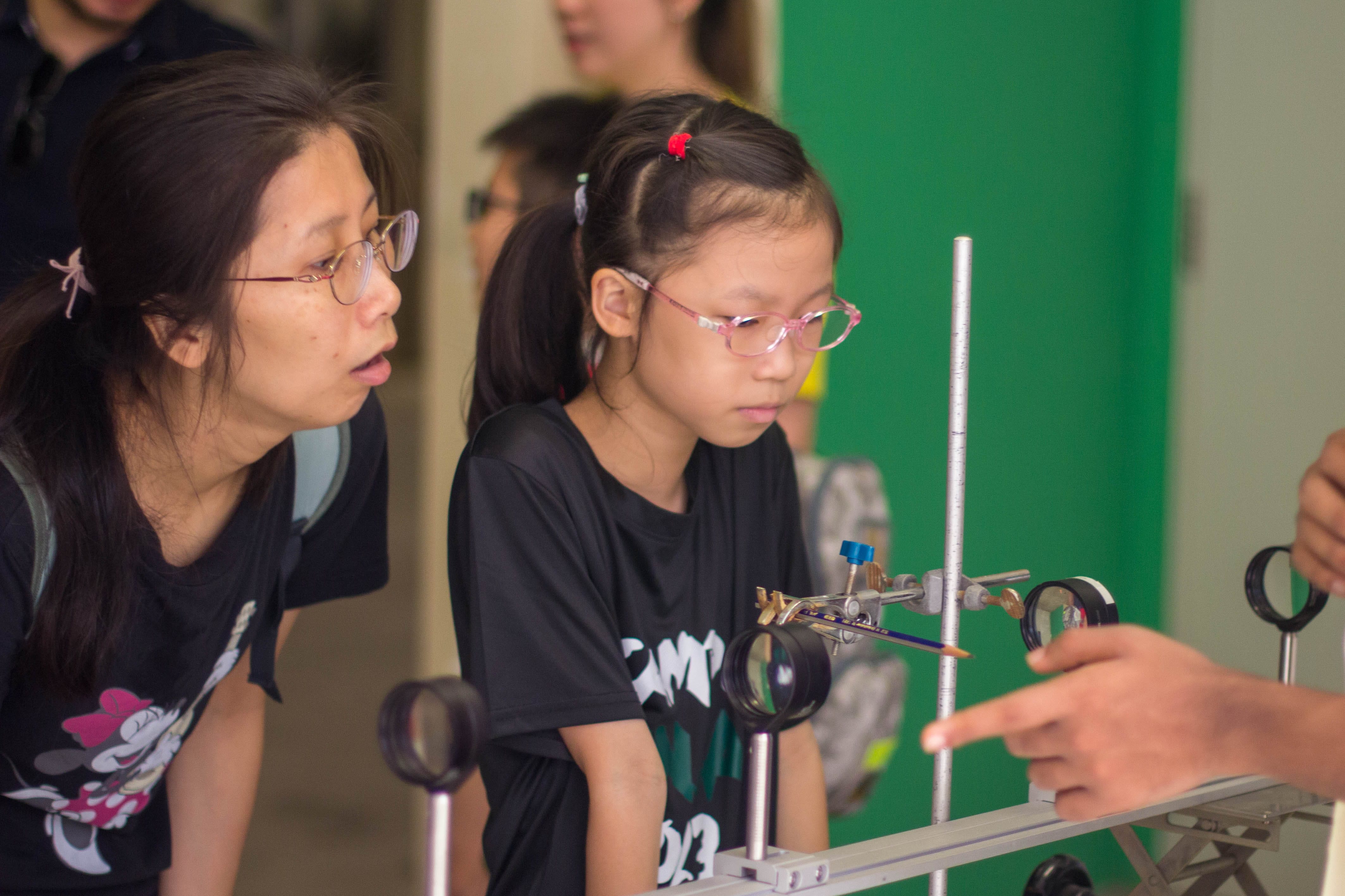A woman and a young girl, both wearing glasses, look closely at a science optical bench with lenses.