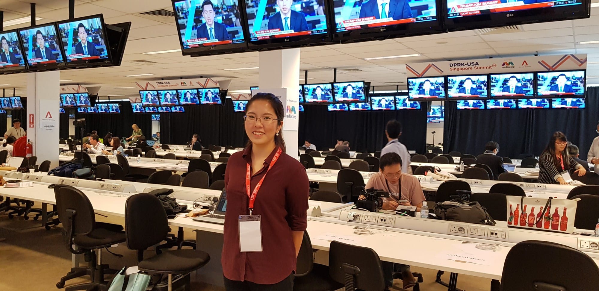 Woman with a red lanyard smiles in a press center with rows of desks and multiple TV screens showing news coverage.