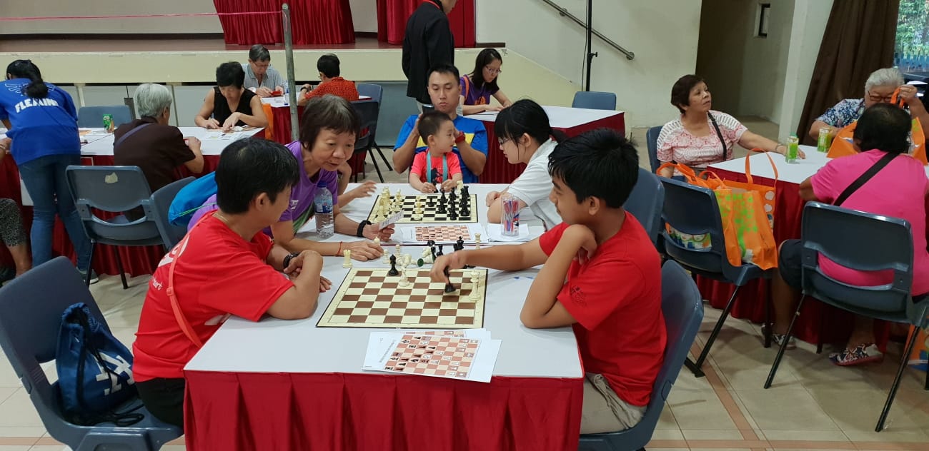 People in a hall sit at tables playing chess; two boys in red shirts face each other at the front table.