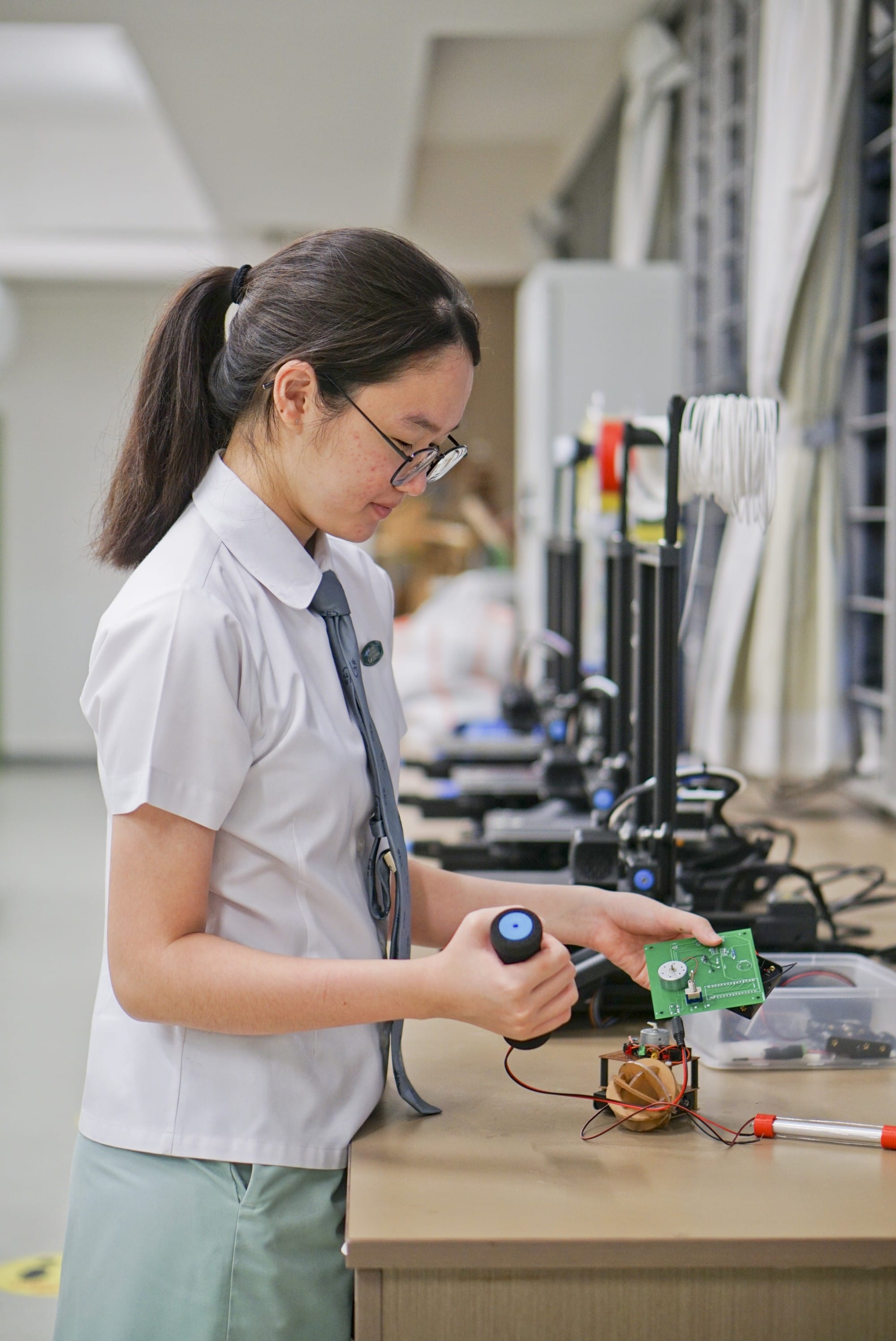 A young female student in uniform holds a circuit board and a wheel while working on a small robot at a desk.