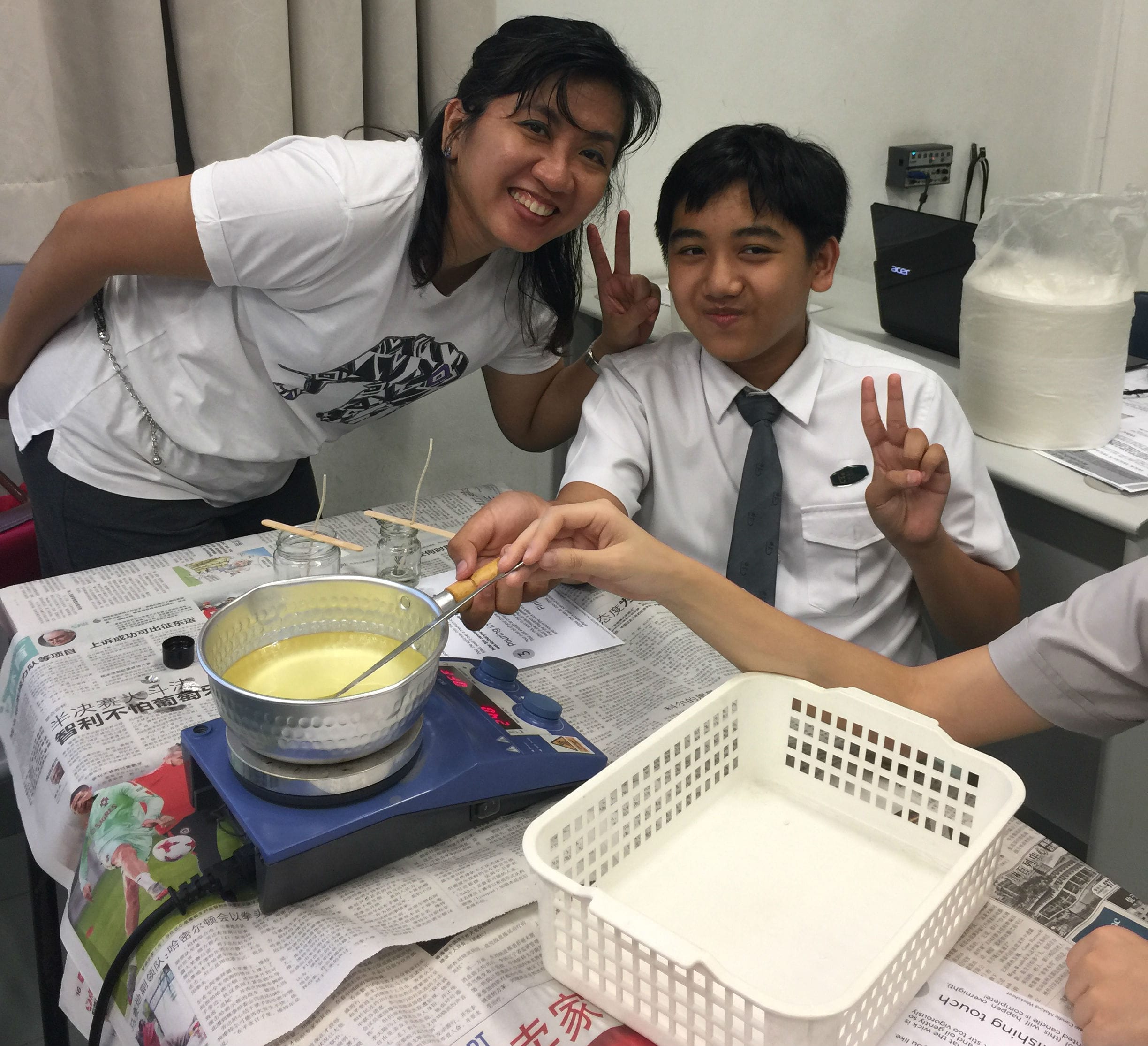 Two people smile and flash peace signs beside a small pot of yellow liquid on a portable stove.