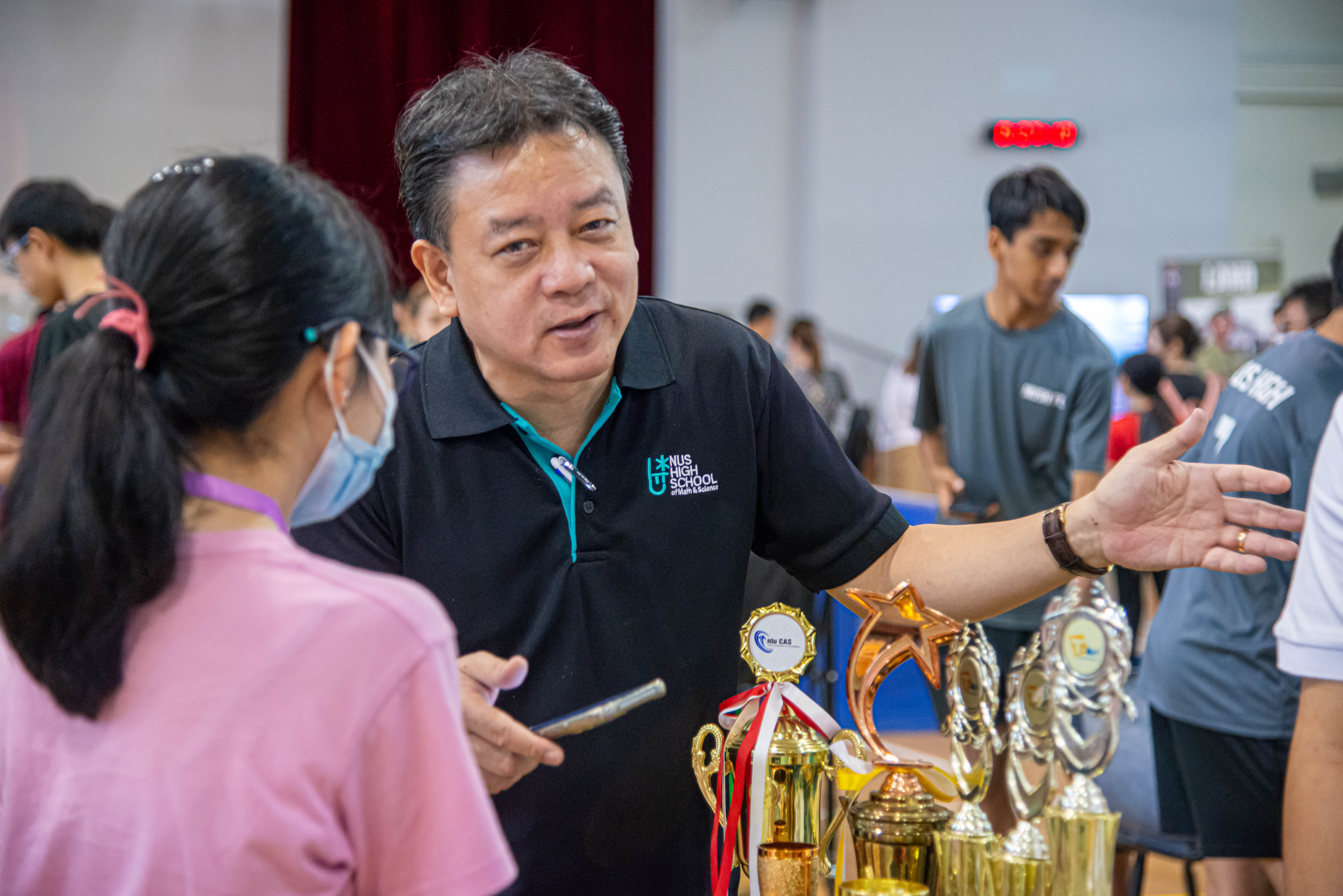 A man in an NUS High School polo shirt gestures toward trophies while speaking to a masked girl in a crowded hall.