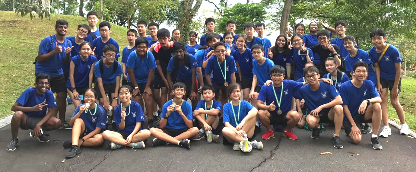 Large group of students in matching blue shirts posing outdoors, some wearing medals, with trees in background.