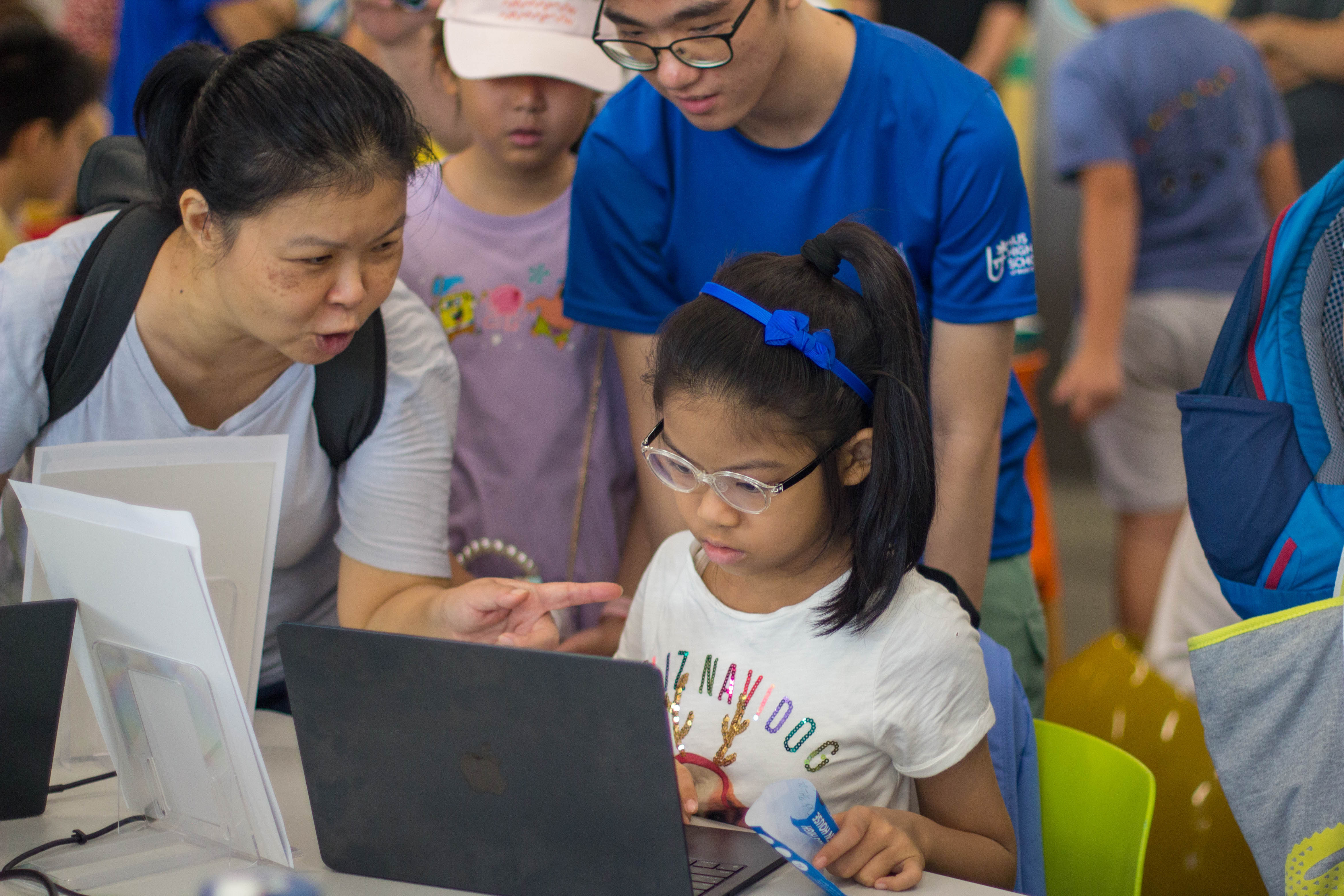 A young girl with glasses uses a laptop while a woman points at the screen and a man in blue looks on.