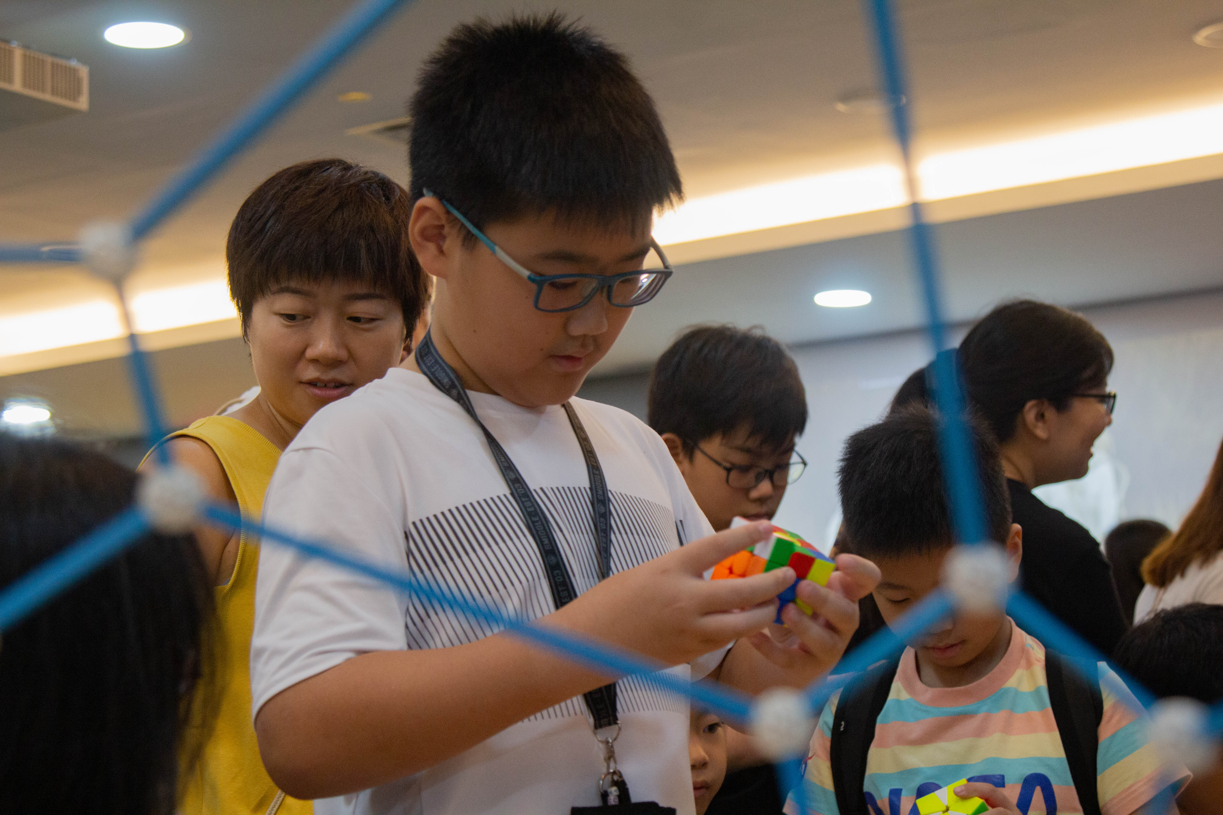 A boy with glasses holds a Rubik's cube while adults and children gather around indoors near a blue geometric structure.