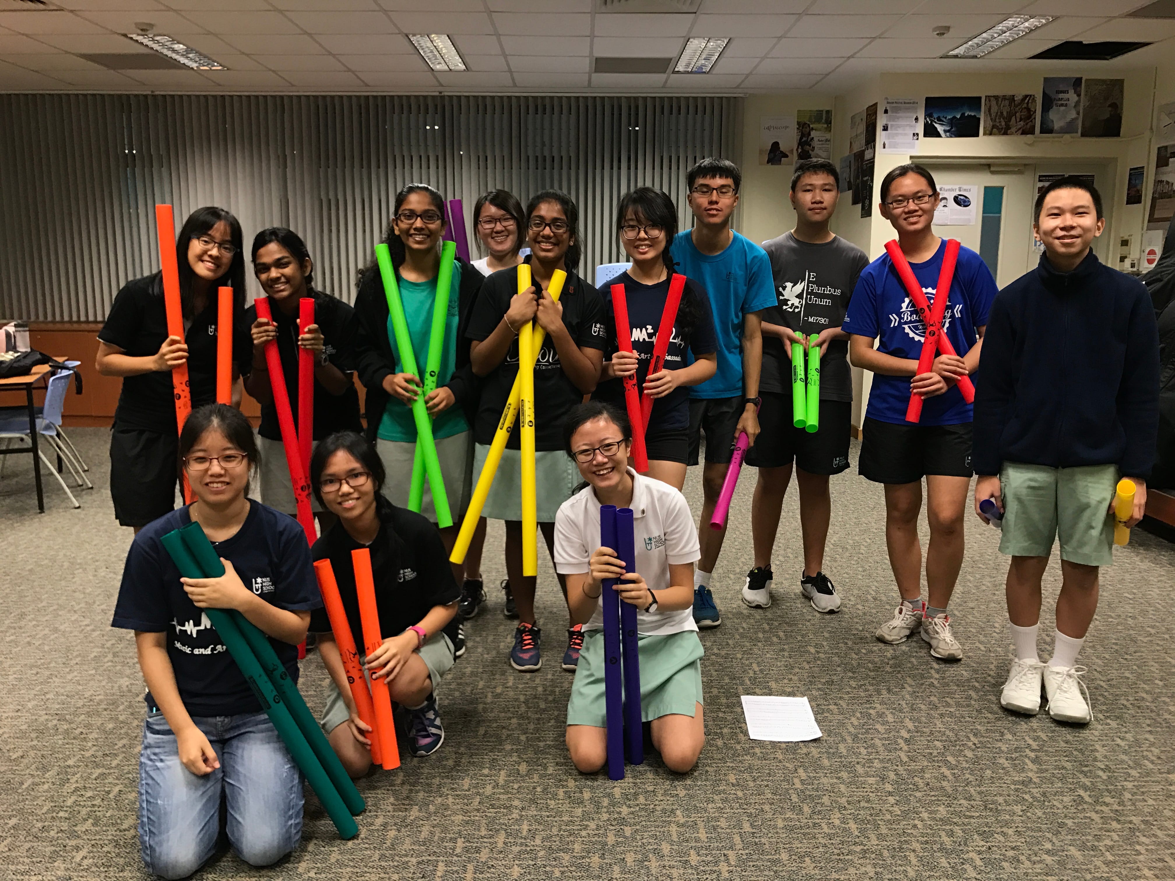 Group of students posing indoors, holding colorful plastic tubes; two kneel in front.
