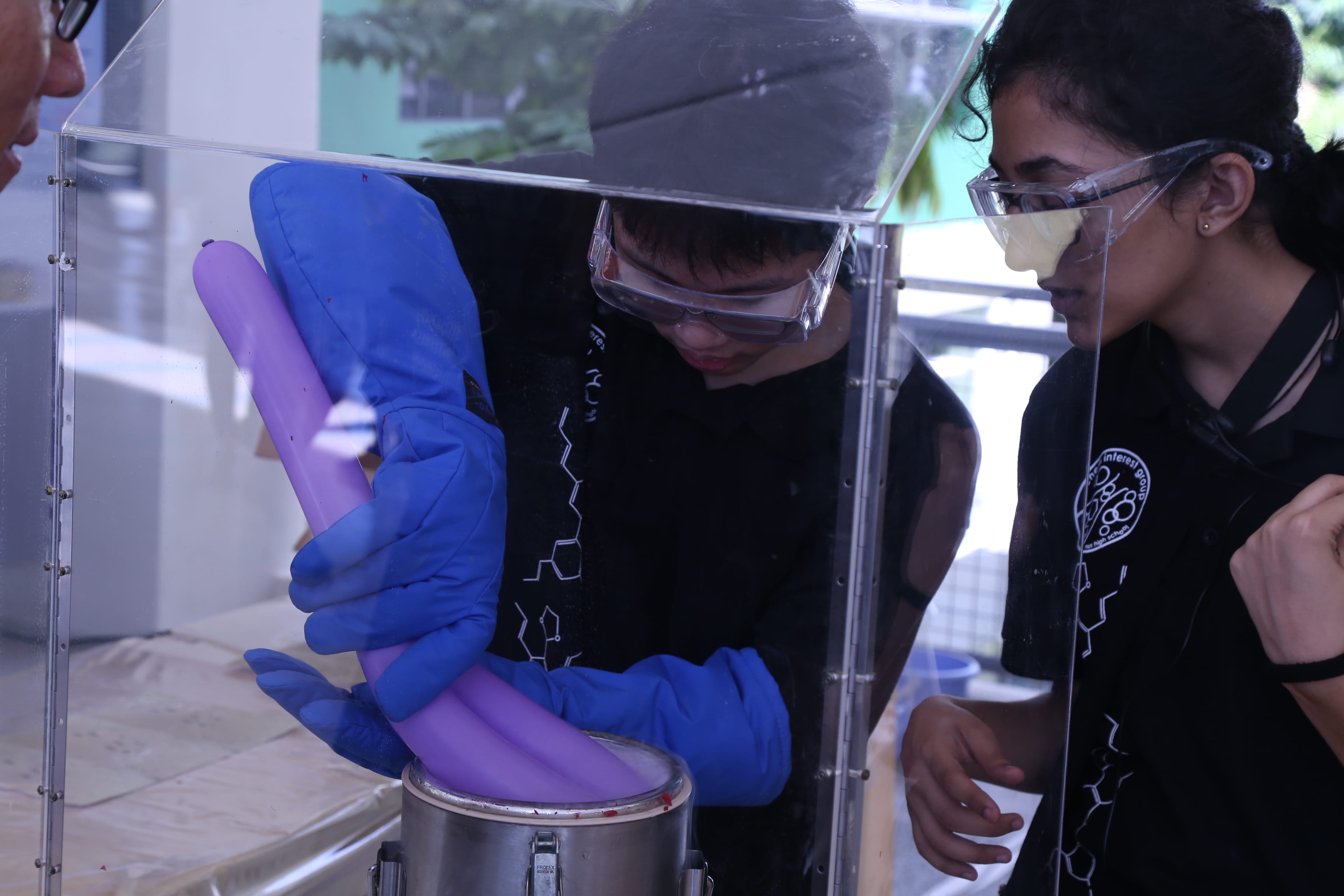 Two people wearing safety goggles and blue gloves handle a purple tube inside a clear enclosure.