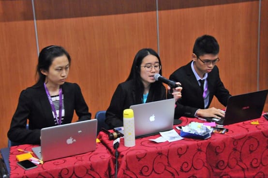 Three people in suits at a red-draped table using laptops; the center person speaks into a microphone.