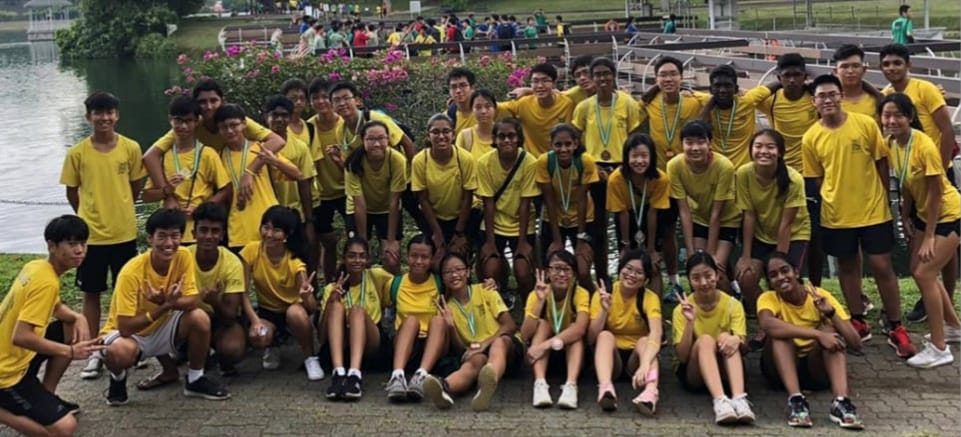 Large group of young people in matching yellow shirts posing outdoors near a waterway with pink flowers behind them.