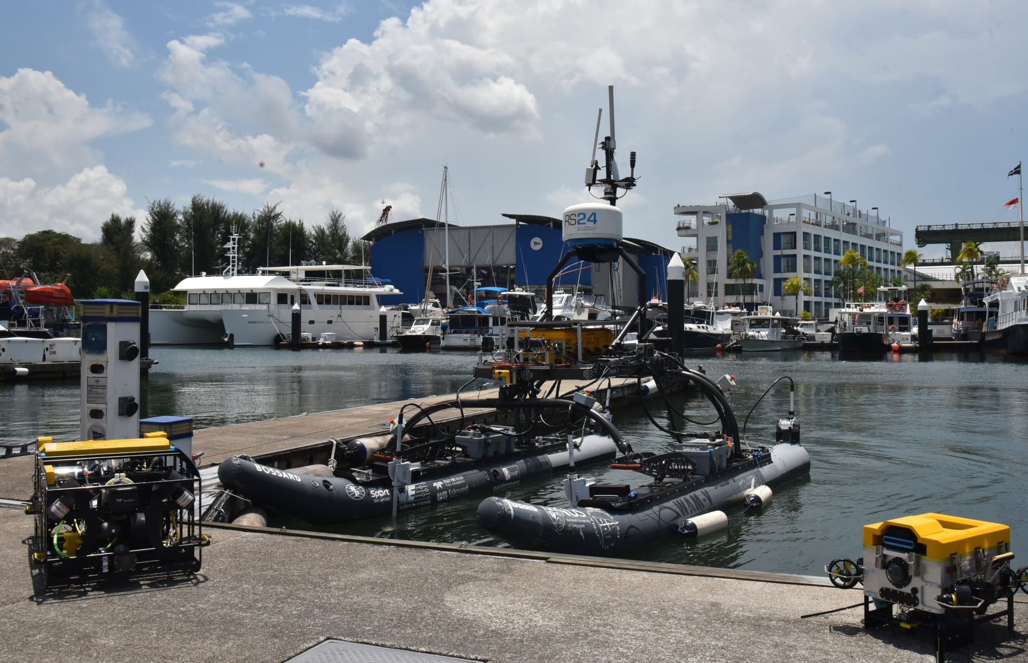 An unmanned surface vessel with sensors docked at a marina, with various boats and buildings in the background.