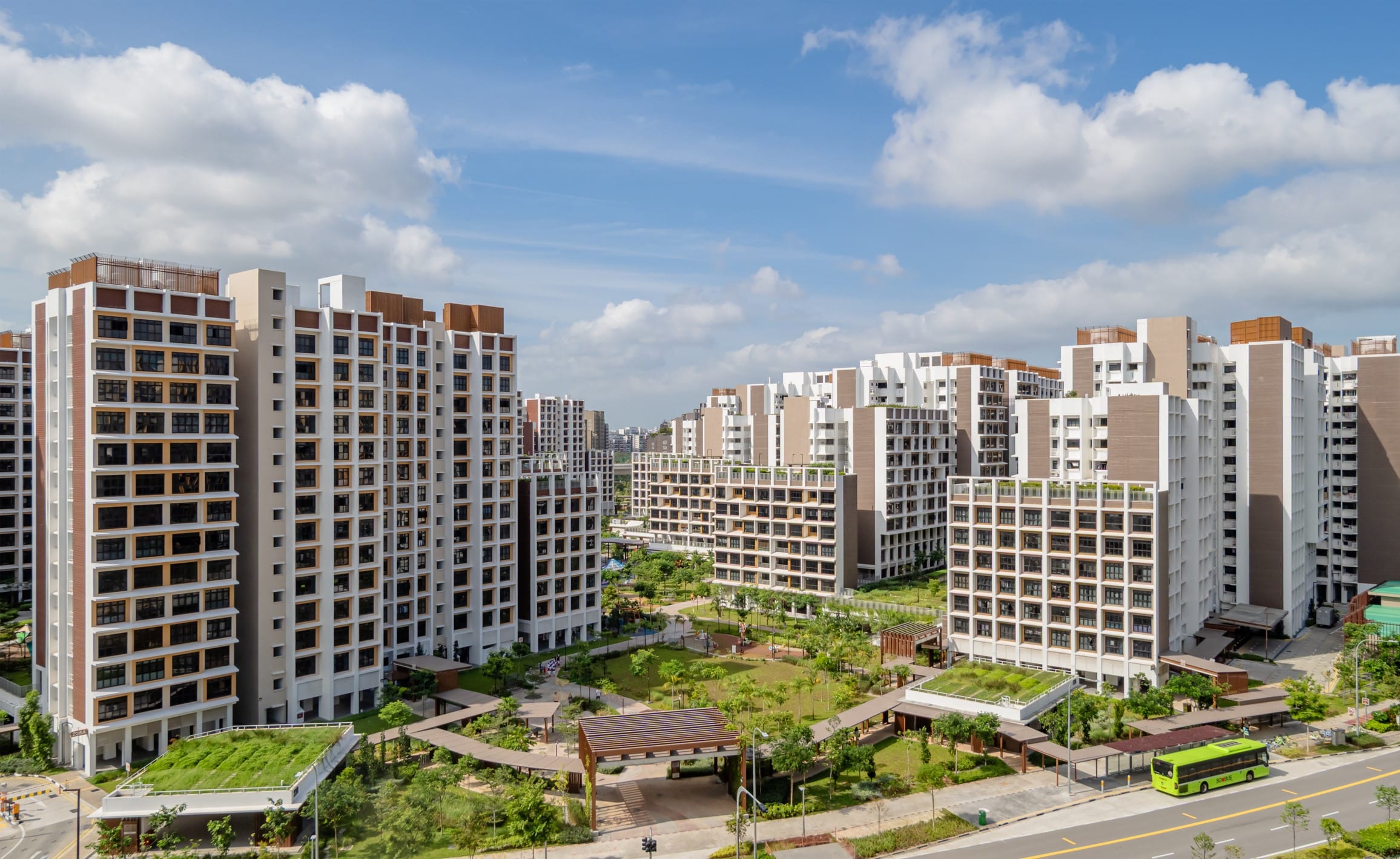 Modern white apartment towers around a green courtyard, with a road and green bus under a blue sky