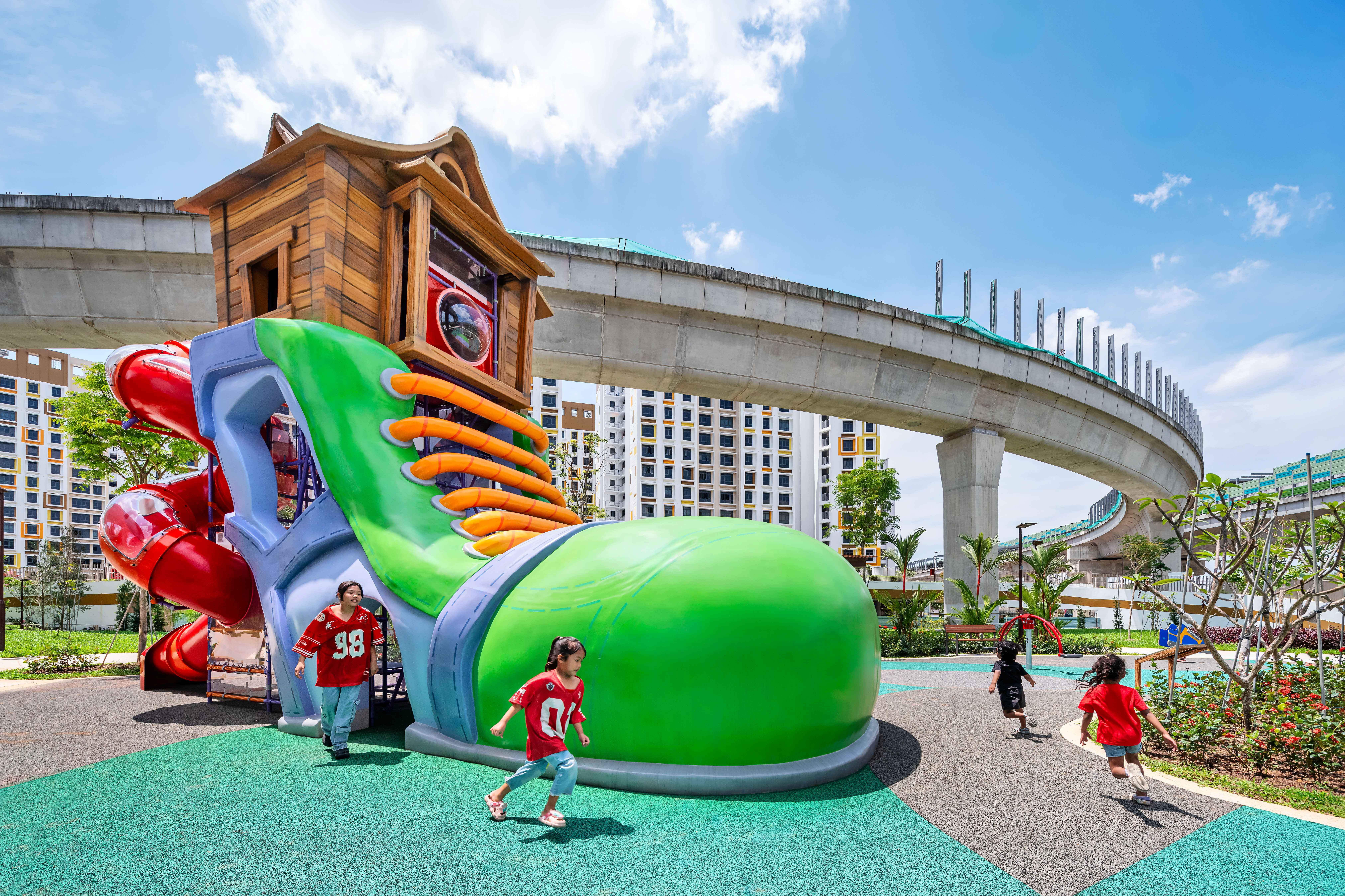 Kids run by a giant green sneaker playground structure with slides, under a curved elevated track and blue sky.