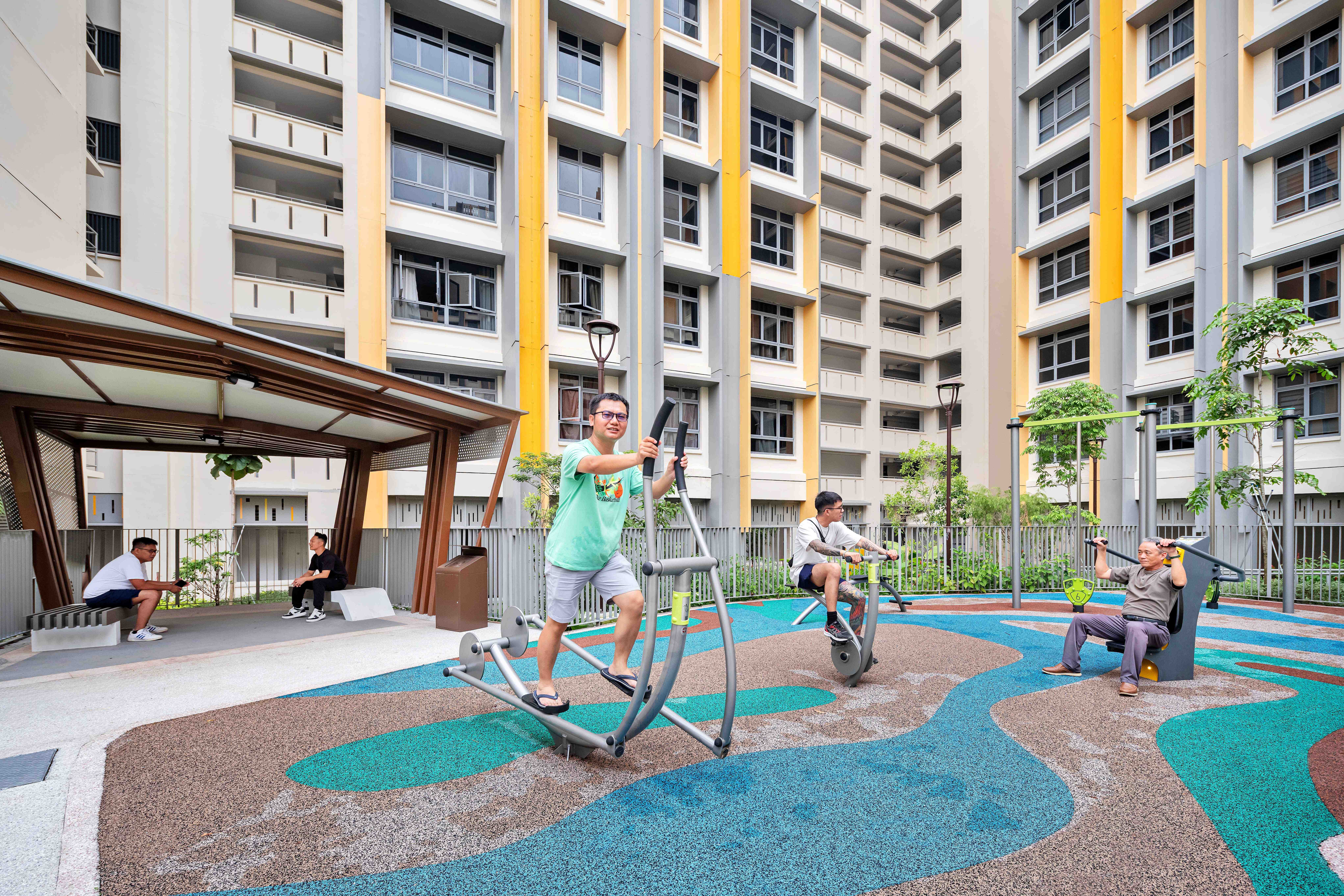 Residents use outdoor gym equipment in a courtyard, with tall apartment blocks and a shaded seating area.
