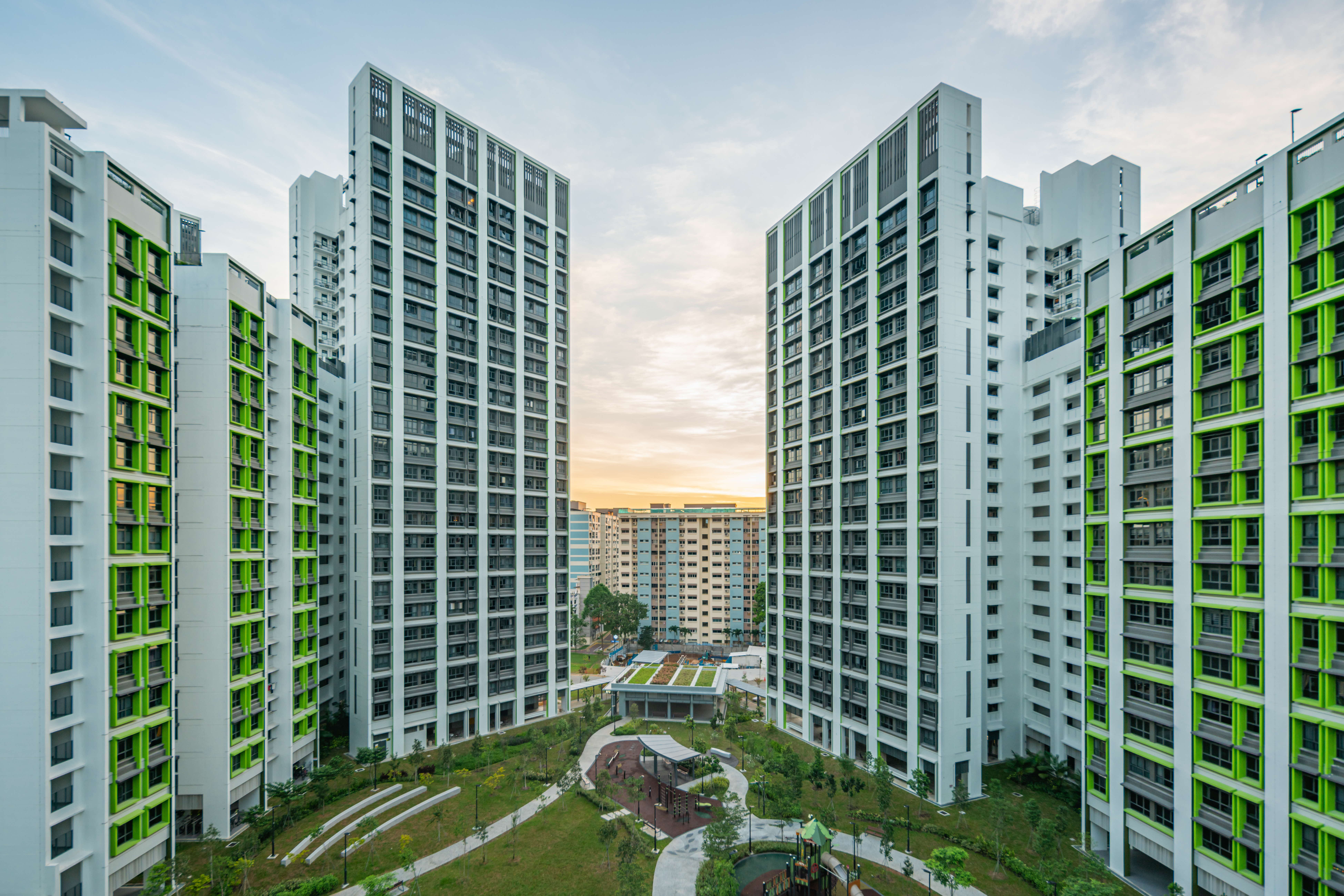 Tall apartment towers with green accents around a landscaped courtyard, pathways, and playground at sunset.