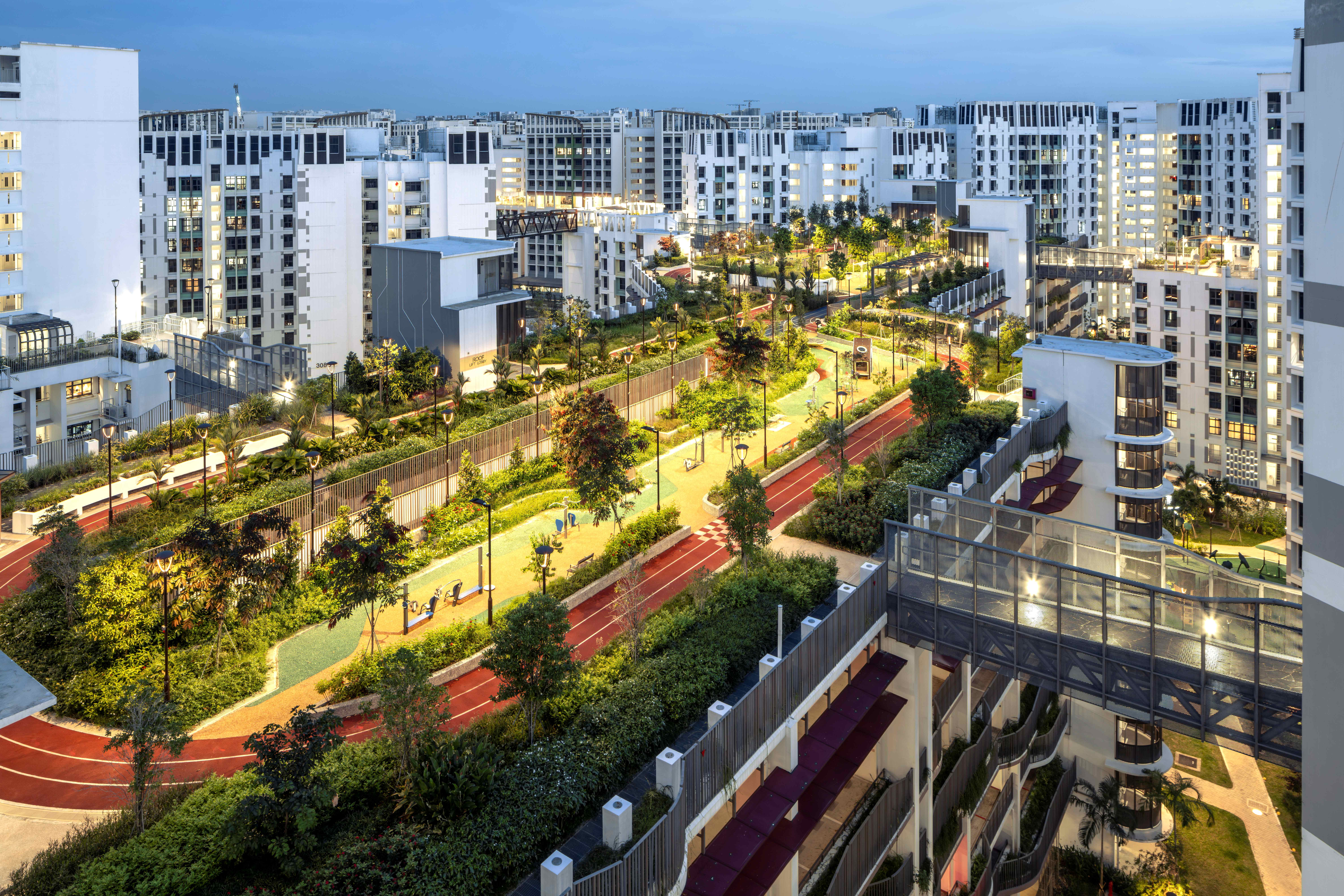 Elevated view of modern apartment towers around a lit park with winding red track, trees, and paths at dusk.